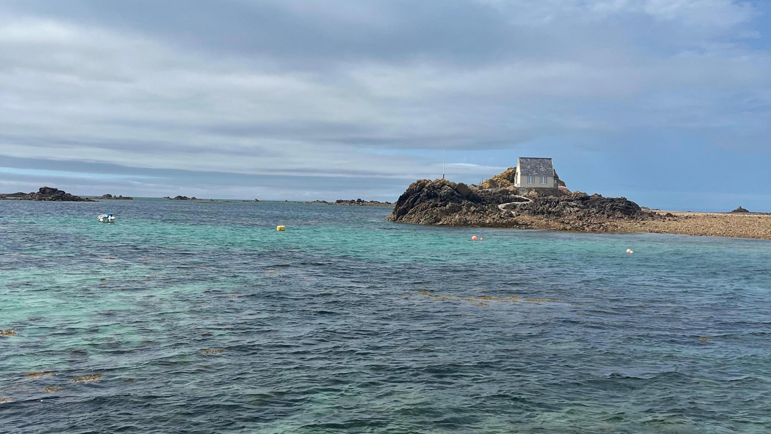 The ocean surrounding a small area of rock with a small house on it. Blue skies with a few grey clouds.