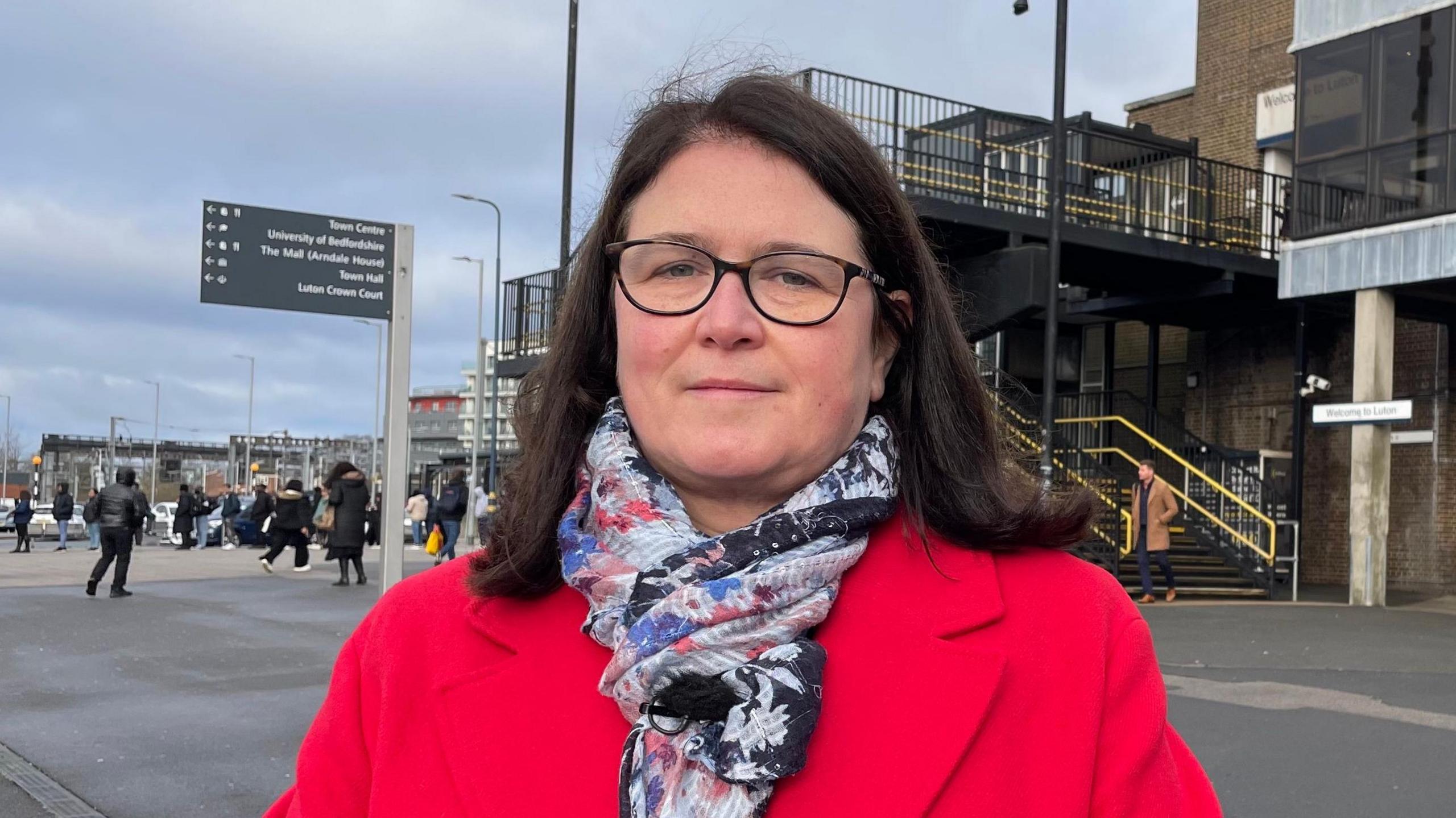 Rachel Hopkins, wearing a bright red overcoat and patterned silk scarf, is standing outside Luton Station looking direct to camera. She is wearing dark rimmed glasses and has brown, shoulder-length hair. Behind her, the station entrance is seen, with pedestrians milling about and some signs.