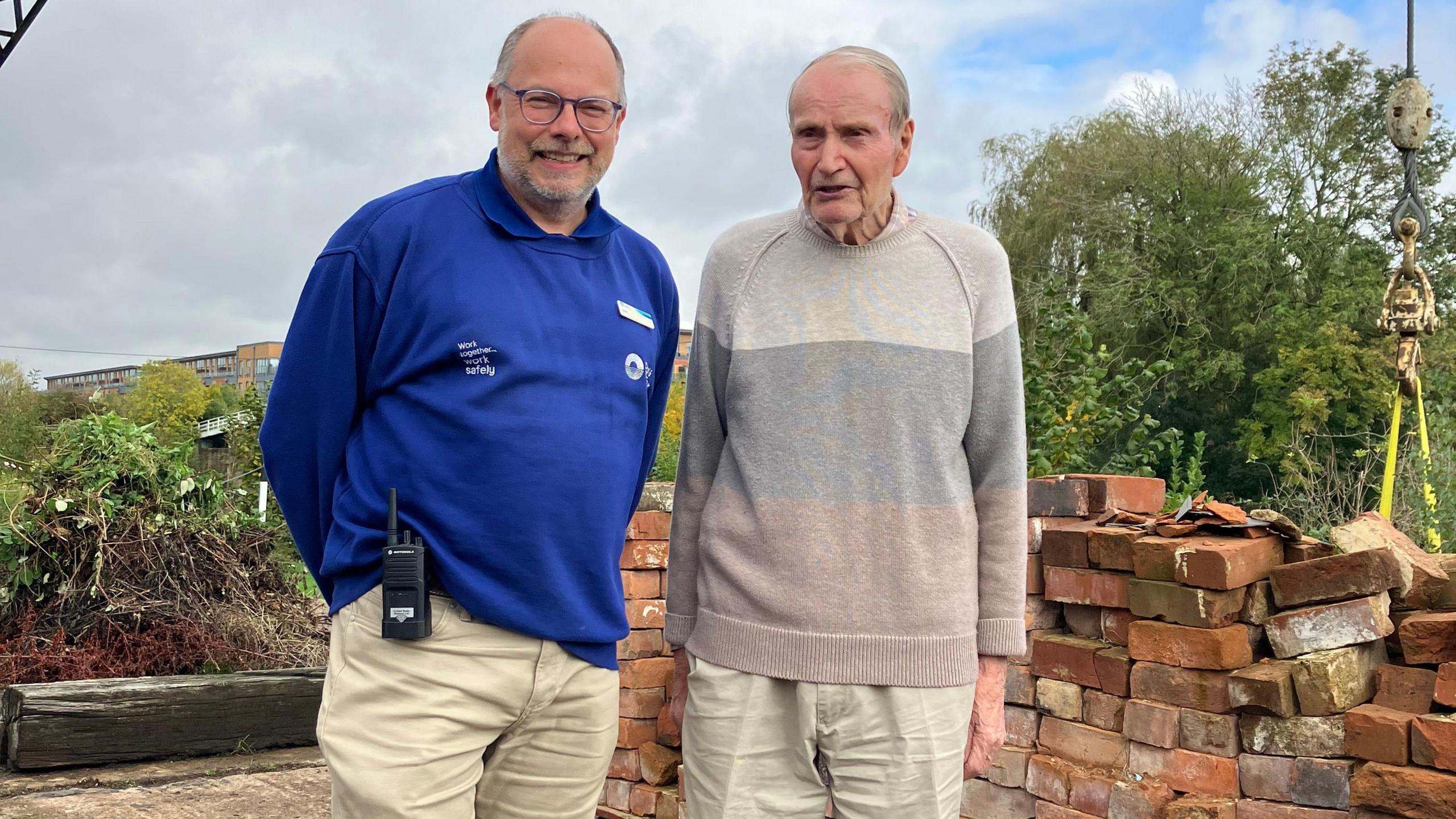 A man wearing a blue jumper stood next to a man wearing a grey jumper in front of a wall and trees at Diglis Locks, with cloudy skies behind them.