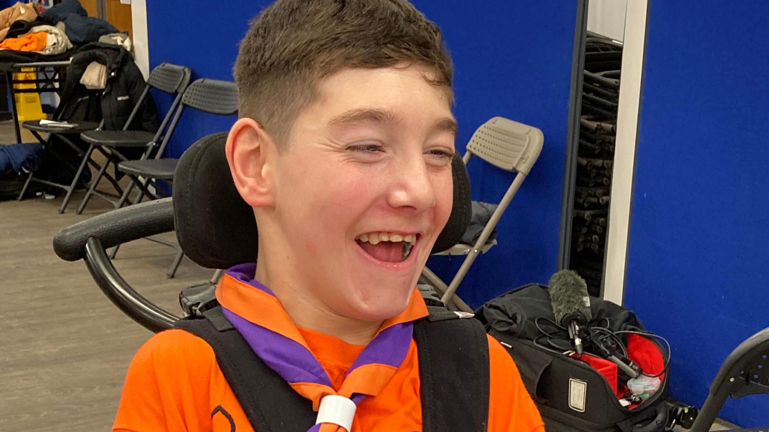 Warwick wears his scout uniform and orange and purple neckerchief as he sits at a computer monitor. He is laughing and looking happy.