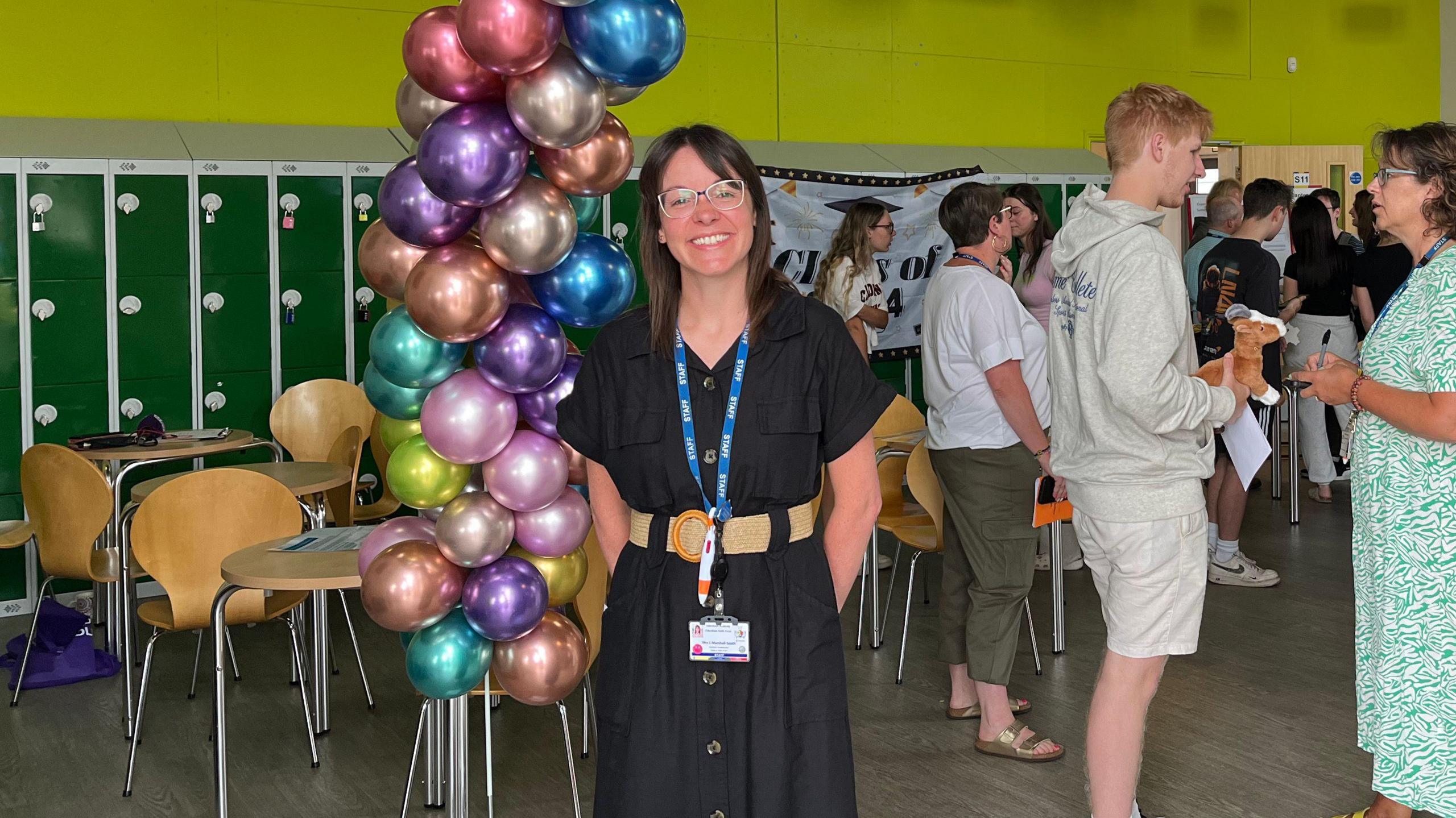 Laura Marshall-Smith has dark brown hair, is wearing a black dress and a tan belt. She is standing next to a balloon arch put up to mark A-level results day at Fakenham Sixth Form. 