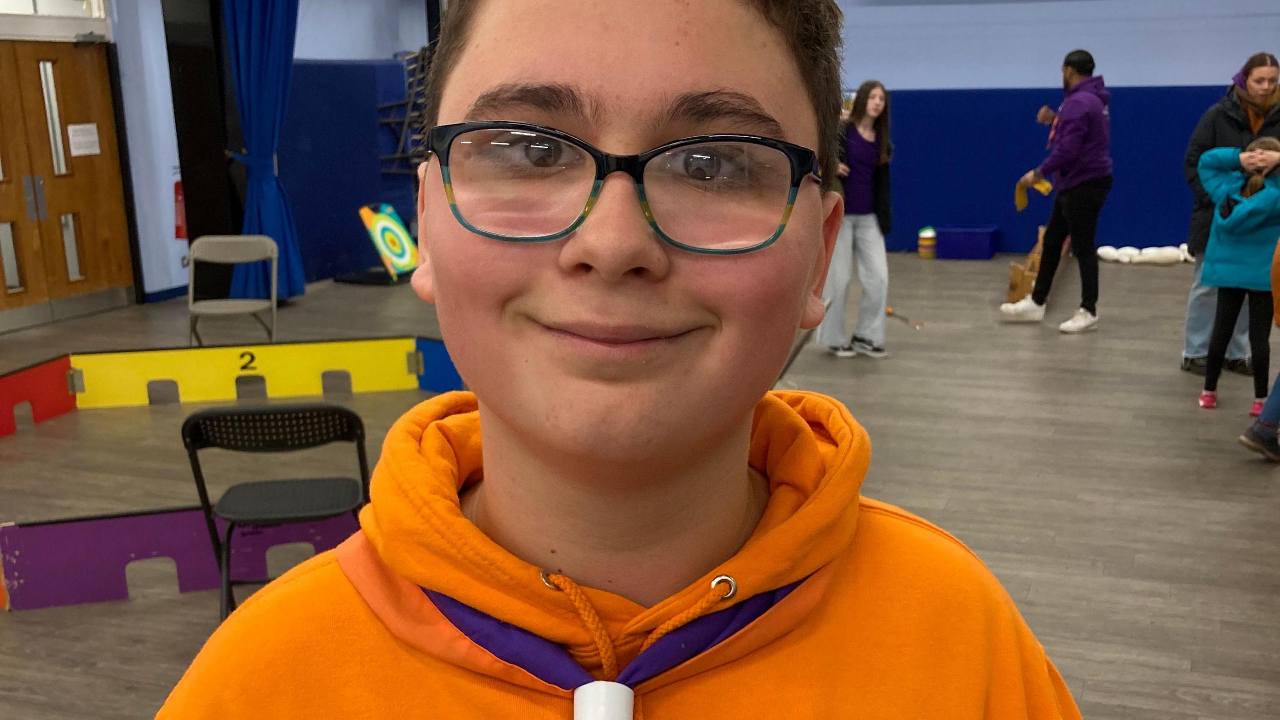 A boy wearing glasses, and an orange scout hoodie with a neckerchief at a scout meeting in a school hall. He is smiling at the cmaera