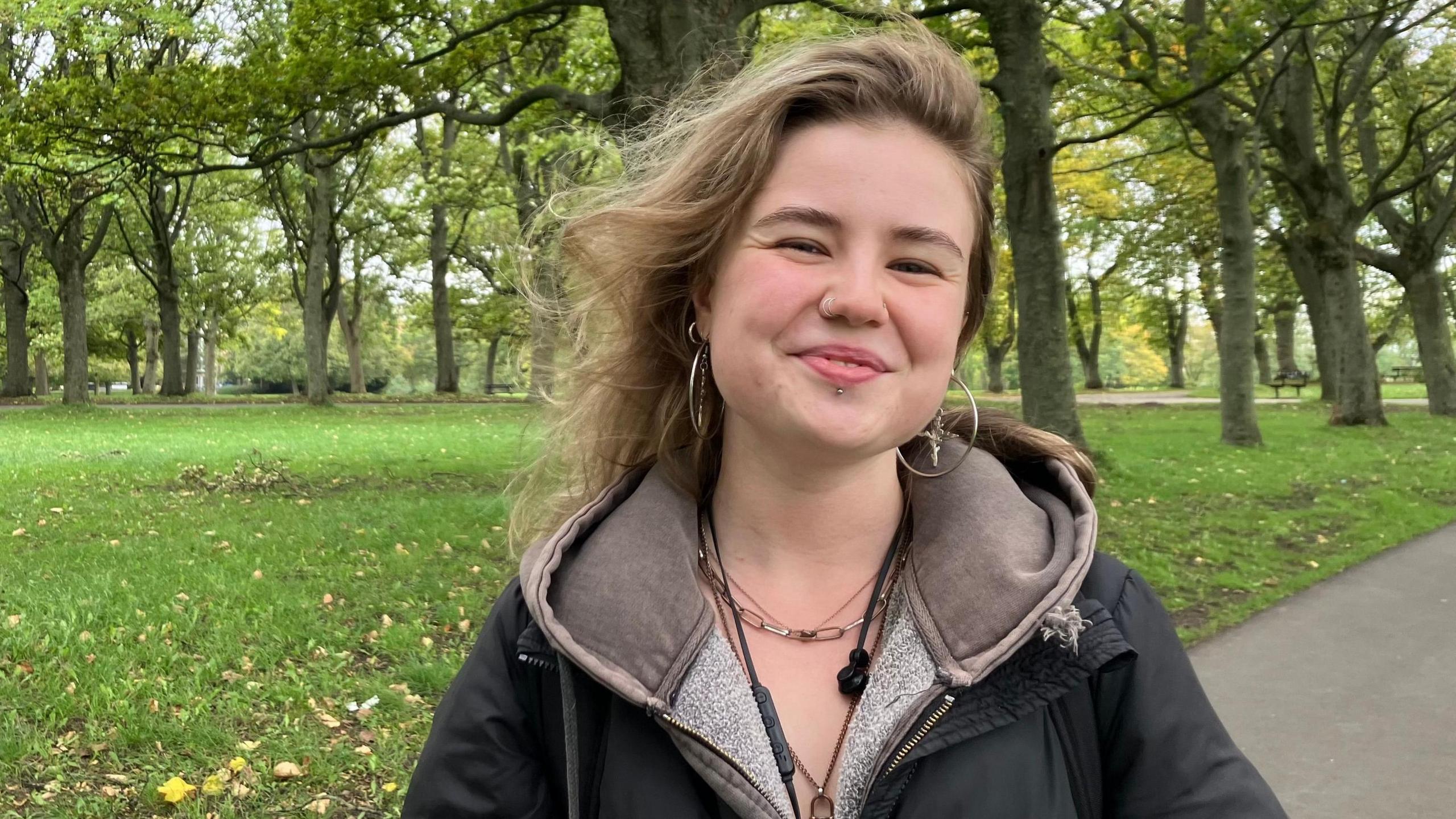 A woman in a park, standing in front of grass and trees. She has long dark blonde hair and is wearing a black jacket and brown hoodie.