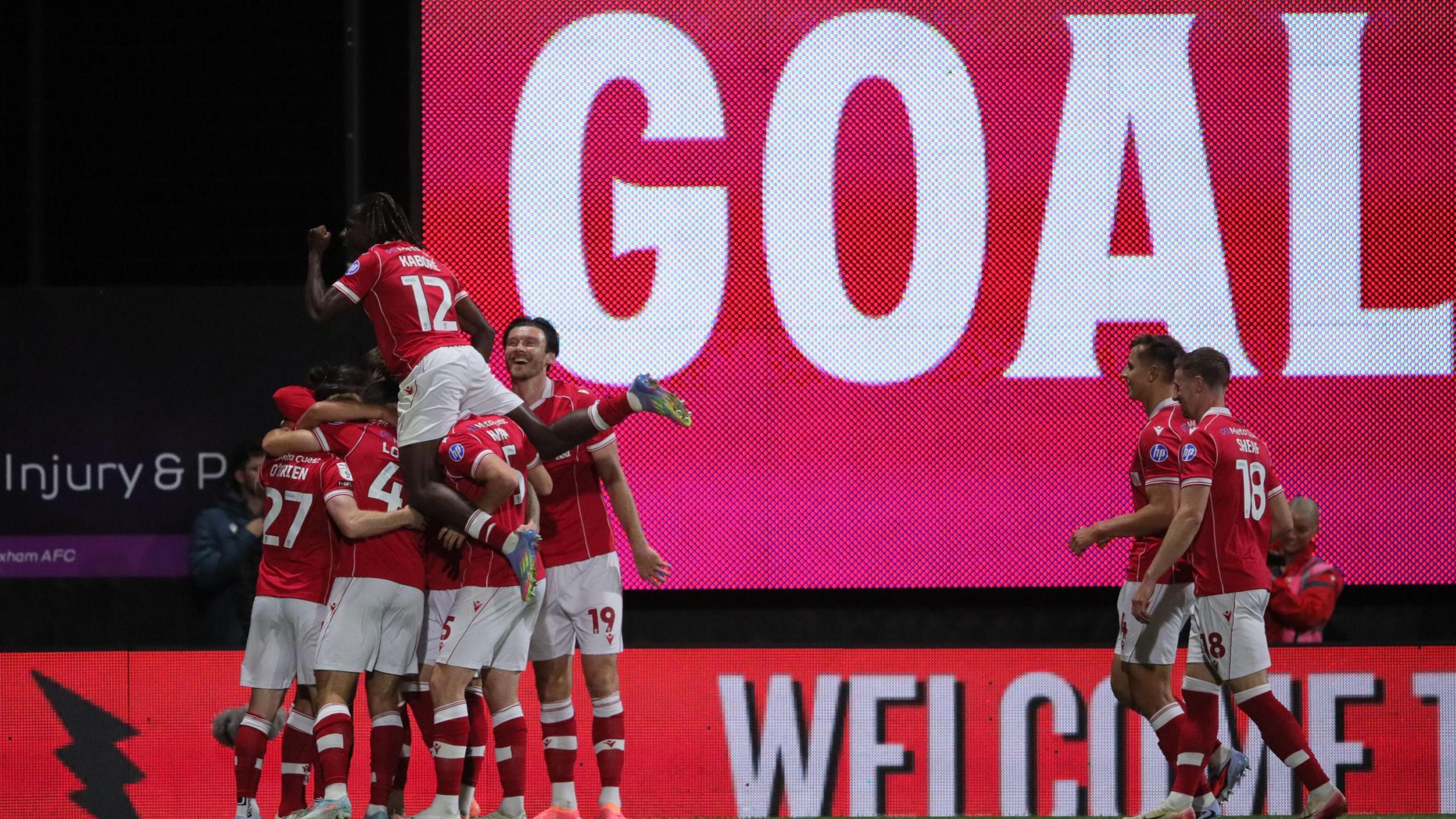 Wrexham players celebrate in a huddle in front of a large scoreboard with the words 'Goal'