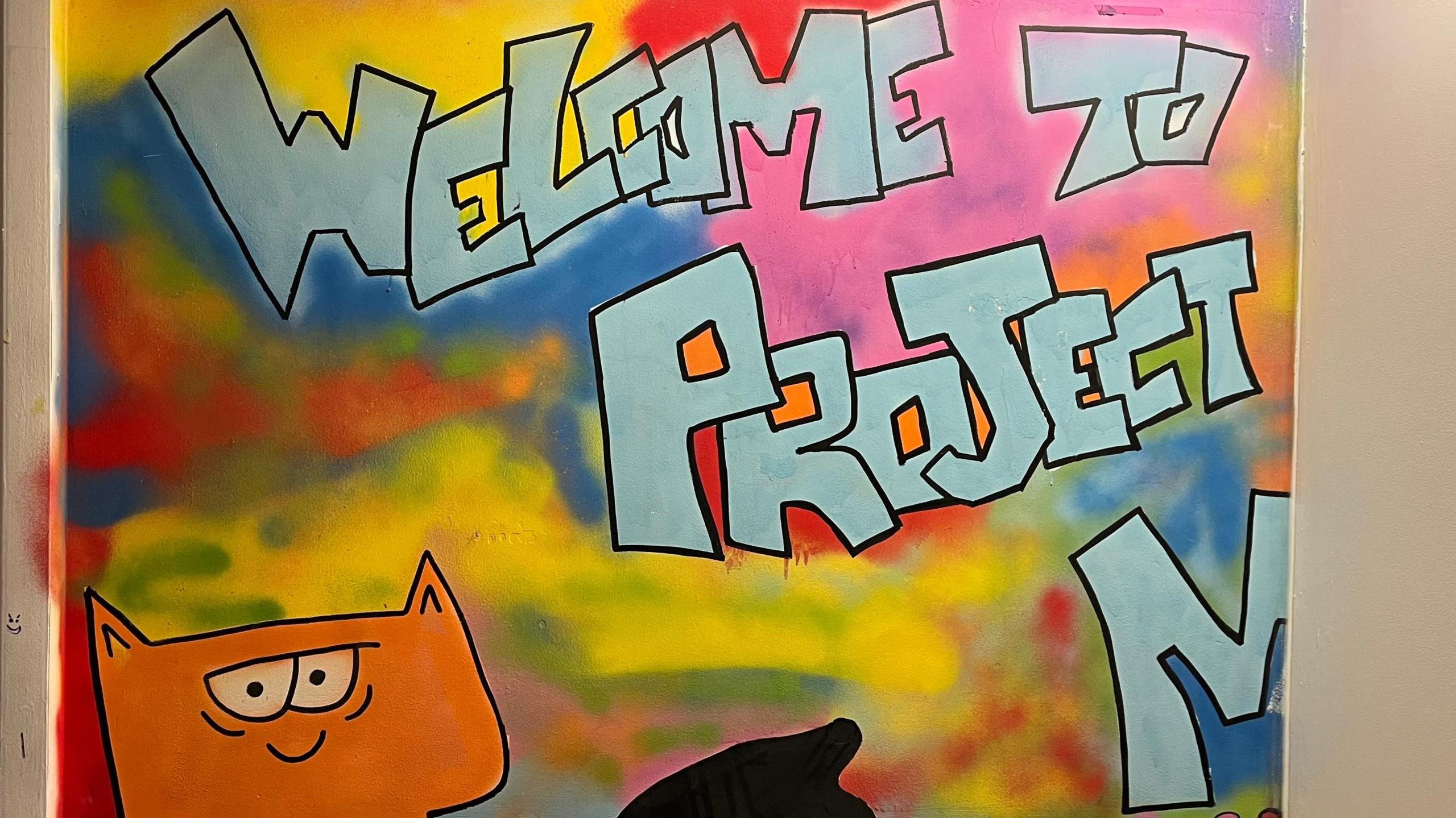 A graffiti inspired wall with "Welcome to Project M" along with a cartoon orange cat on it. 
