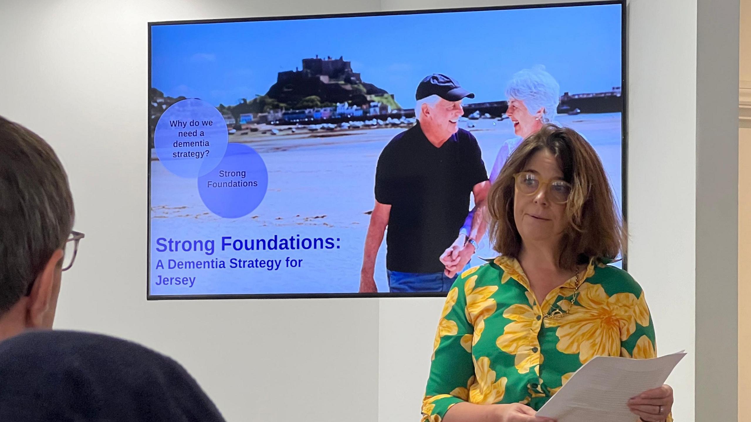 A woman in a floral dress gives a speech in front of a screen which shows the cover image of the Dementia Strategy