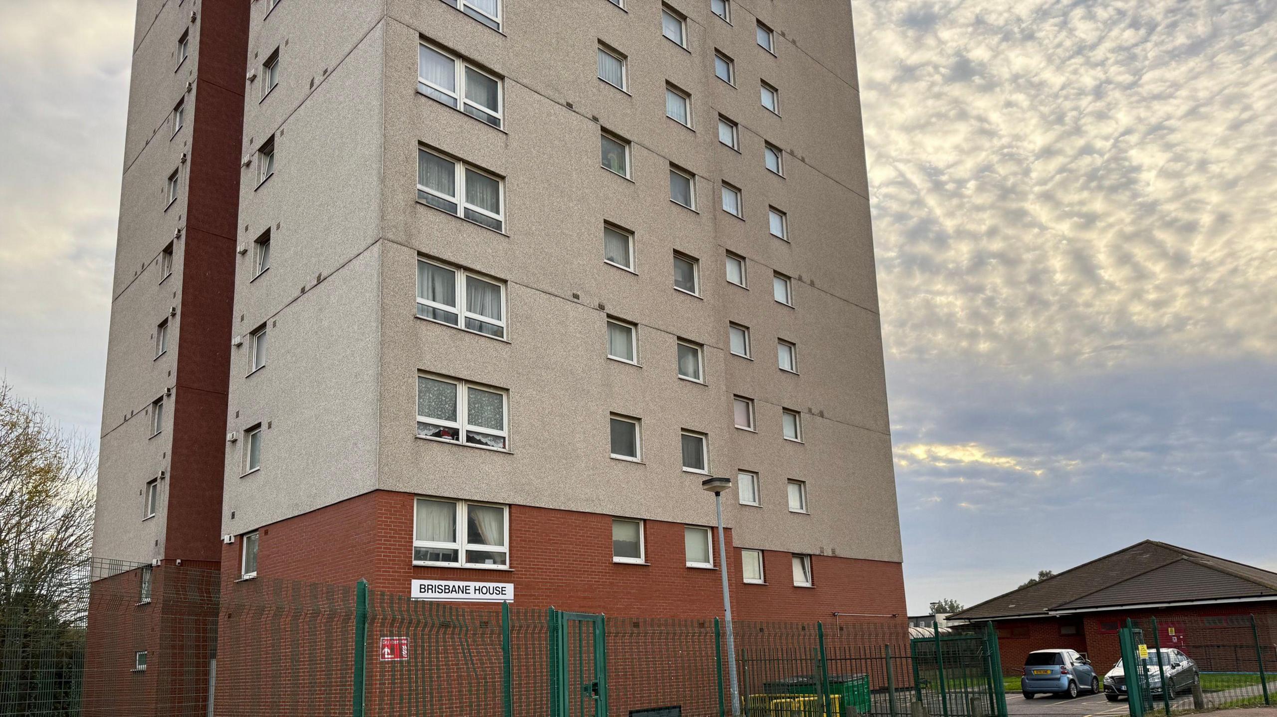 A close up of the tower block, showing the first 8 floors. It is made of grey concrete with red brick on the ground floor. A white sign says Brisbane House on the bottom left of the building. A green metal see-through fence is in the foreground. The sky is bright with dappled clouds.