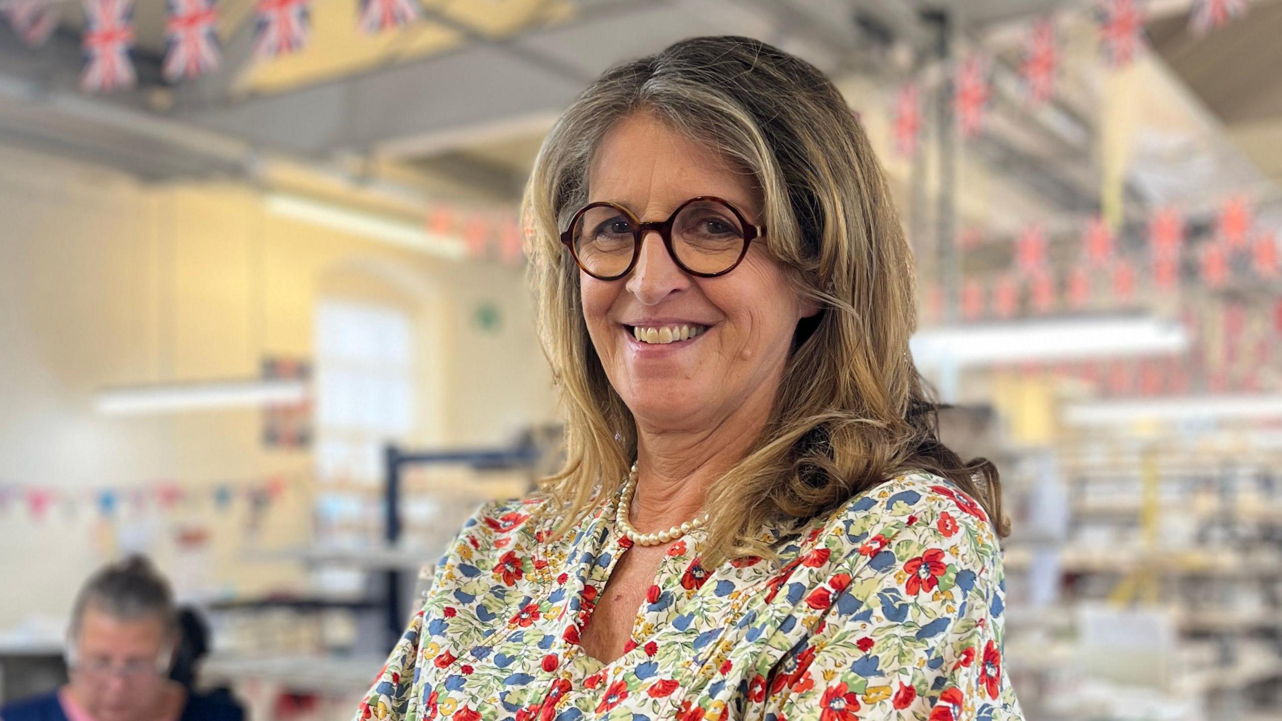 Emma Bridgewater - a woman wearing a patterned dress, glasses and a pearl necklace - is standing with her arms folded. She is smiling for the camera. Behind her is a factory with union jack bunting.