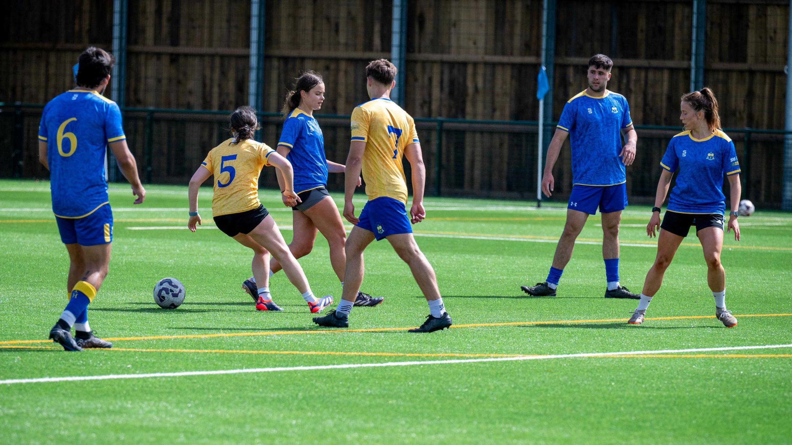 A group of male and female footballers in blue and yellow kits play on the new bright green 3G pitch at the University of Bath