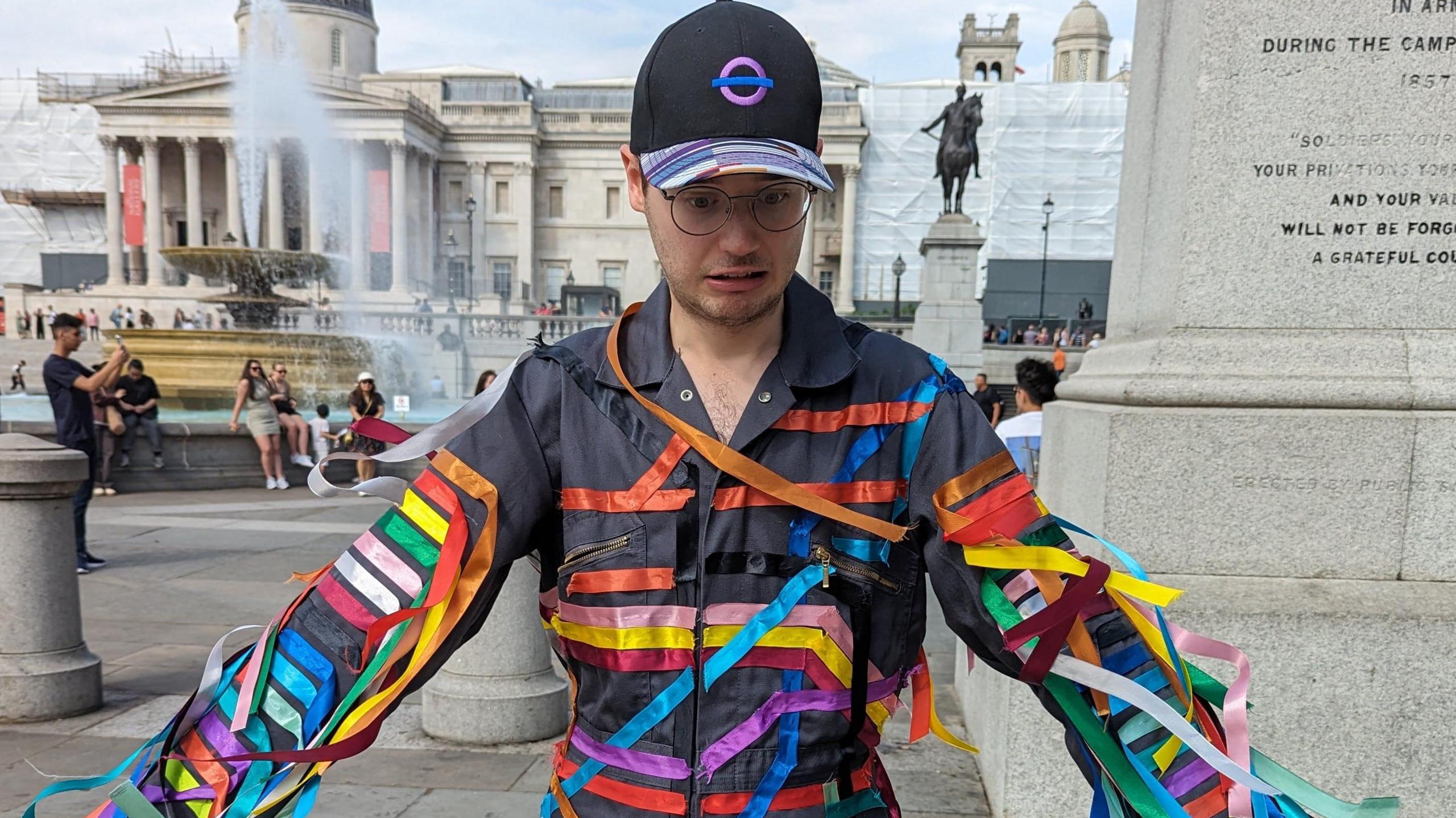 Tom in a Tube line inspired outfit and an Elizabeth line cap. He stands outside Trafalgar Square.