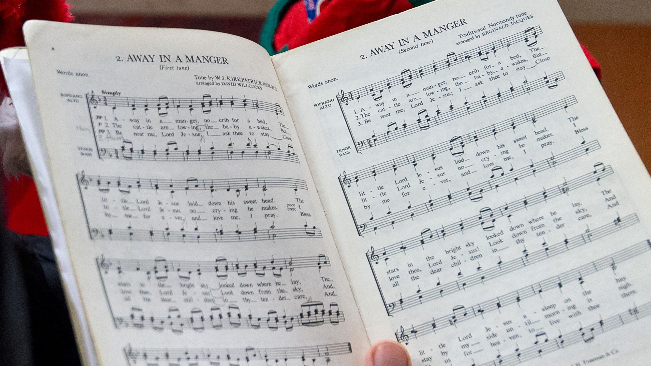 An open music book containing the musical score of Away In A Manger. A person's thumb is placed at the bottom of the book as they hold it up. 