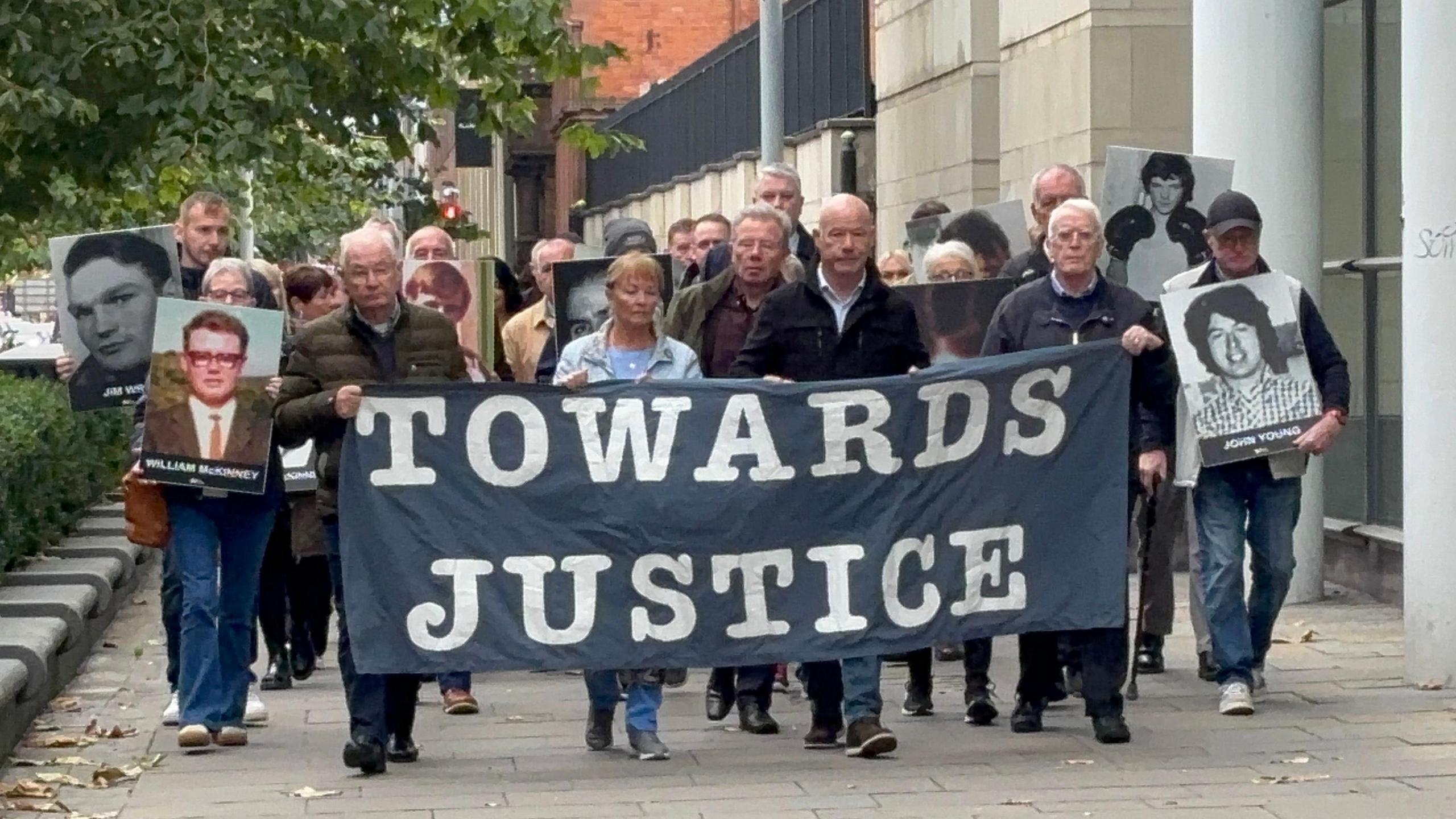 A group of around twenty people walk together outside a court building. They are holding a banner that reads towards justice, as well as posters of four individuals. Leaves can be seen on the ground and a number of trees are visible behind the group.