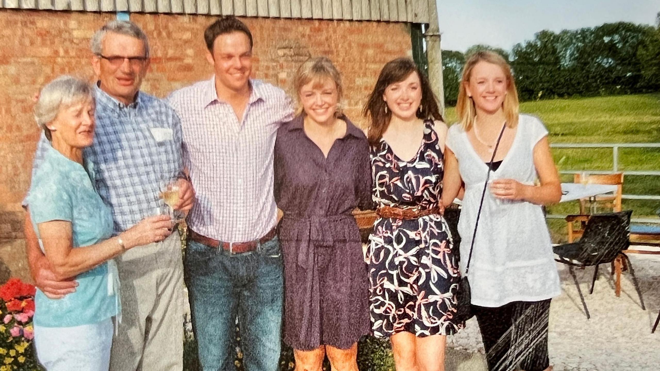 Sheena and Michael Read stand with their four grown-up children in front of a red-brick wall in a farm yard. They wear summer clothes and a table and chairs can be seen to one side. In the background is a green paddock and trees.