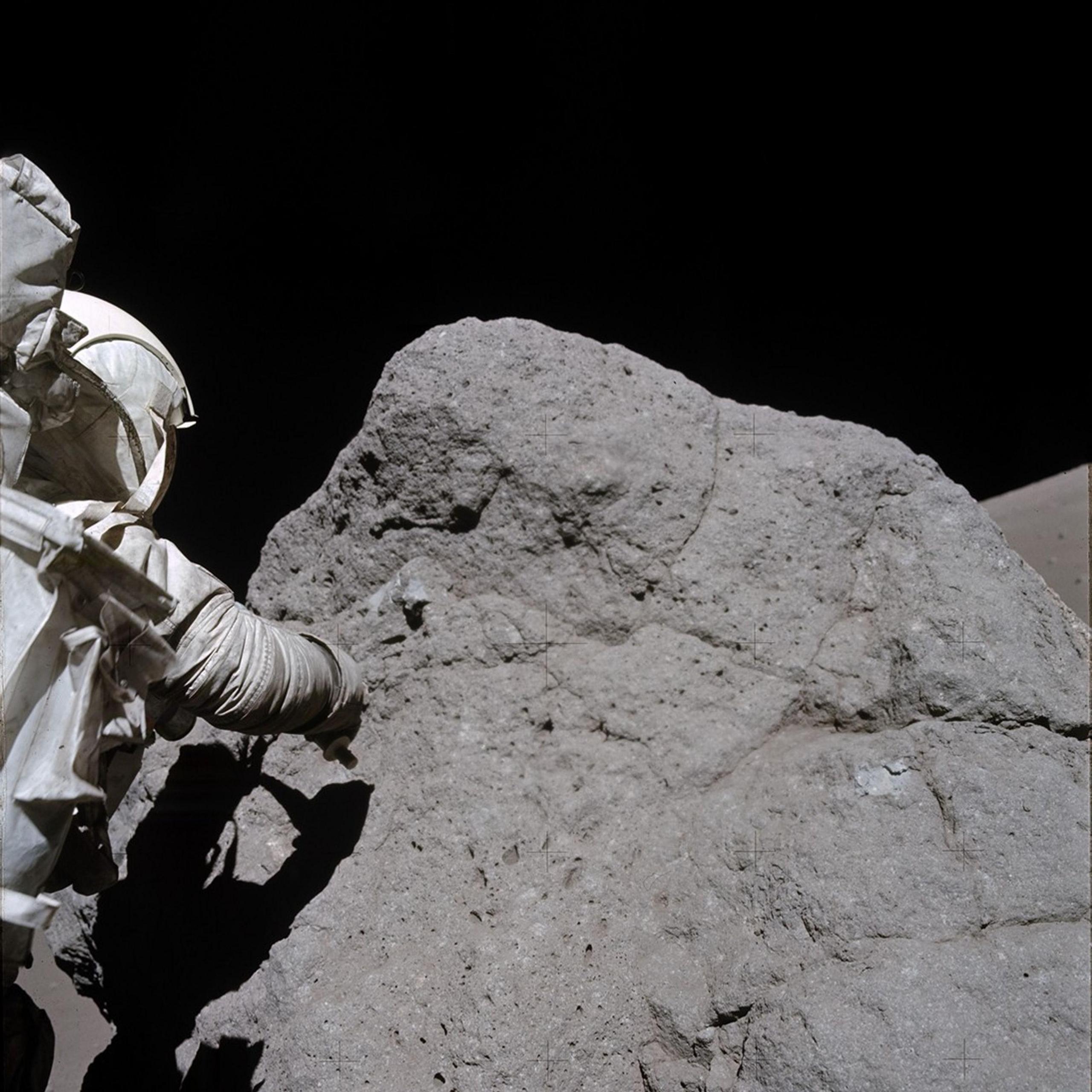 The Apollo astronaut Harrison Schmitt, from Apollo 17,  is collecting samples  rock on the Moon. The rock is slightly taller than him and about 3 times as wide. He is on the left of the photo with his back to the camera, and is wearing a white NASA space suit as he tries to collect the sample using his right arm. His body is creating a shadow on the large dark grey rock. At the top of the photo you can see the blackness of space. 

