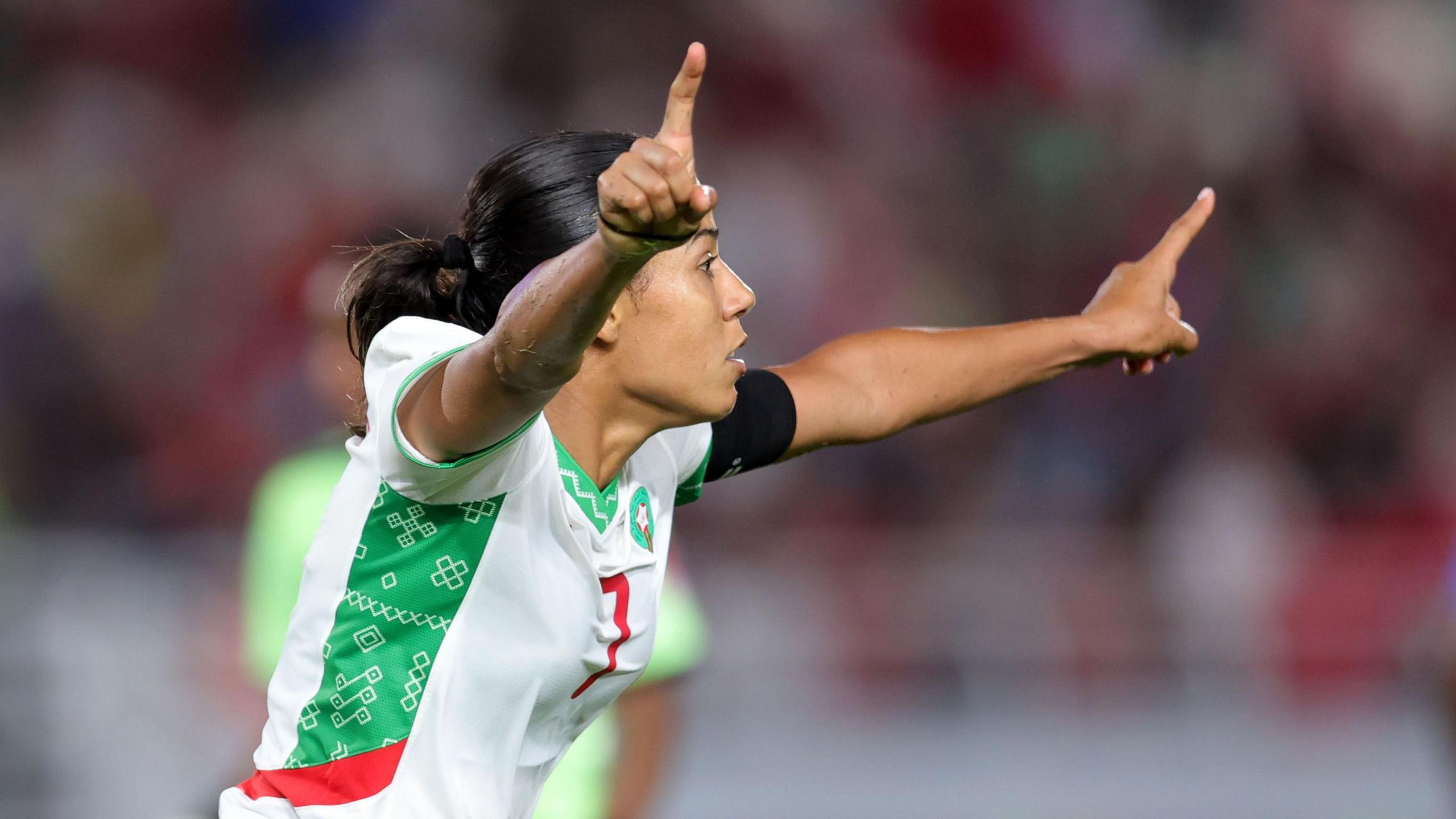 Ghizlane Chebbak, wearing a white Morocco jersey with green and red trim, raises her arms and points with her index fingers as she celebrates