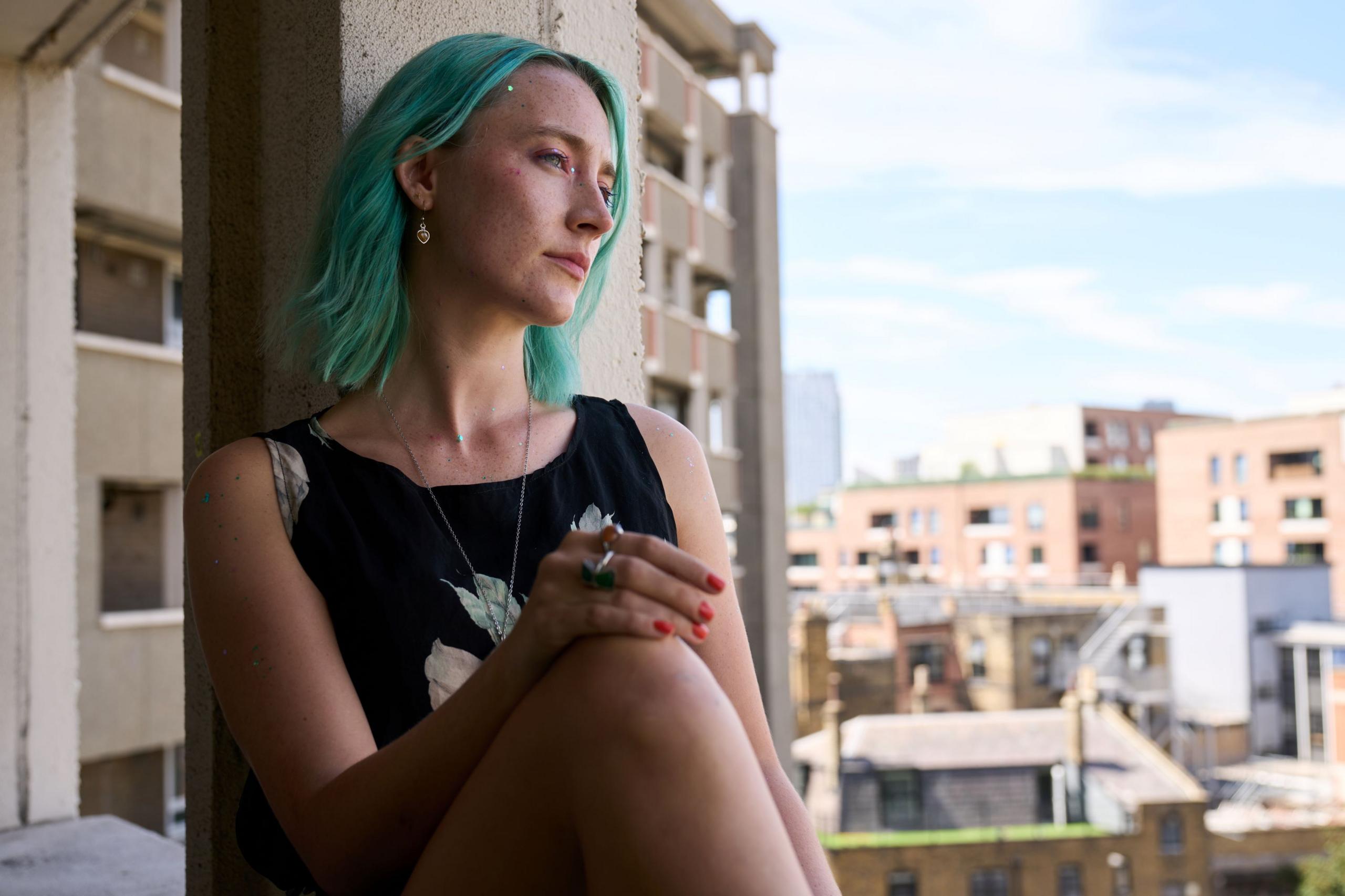 Saoirse, as Rona, with green hair, sitting at the window of a flat in London