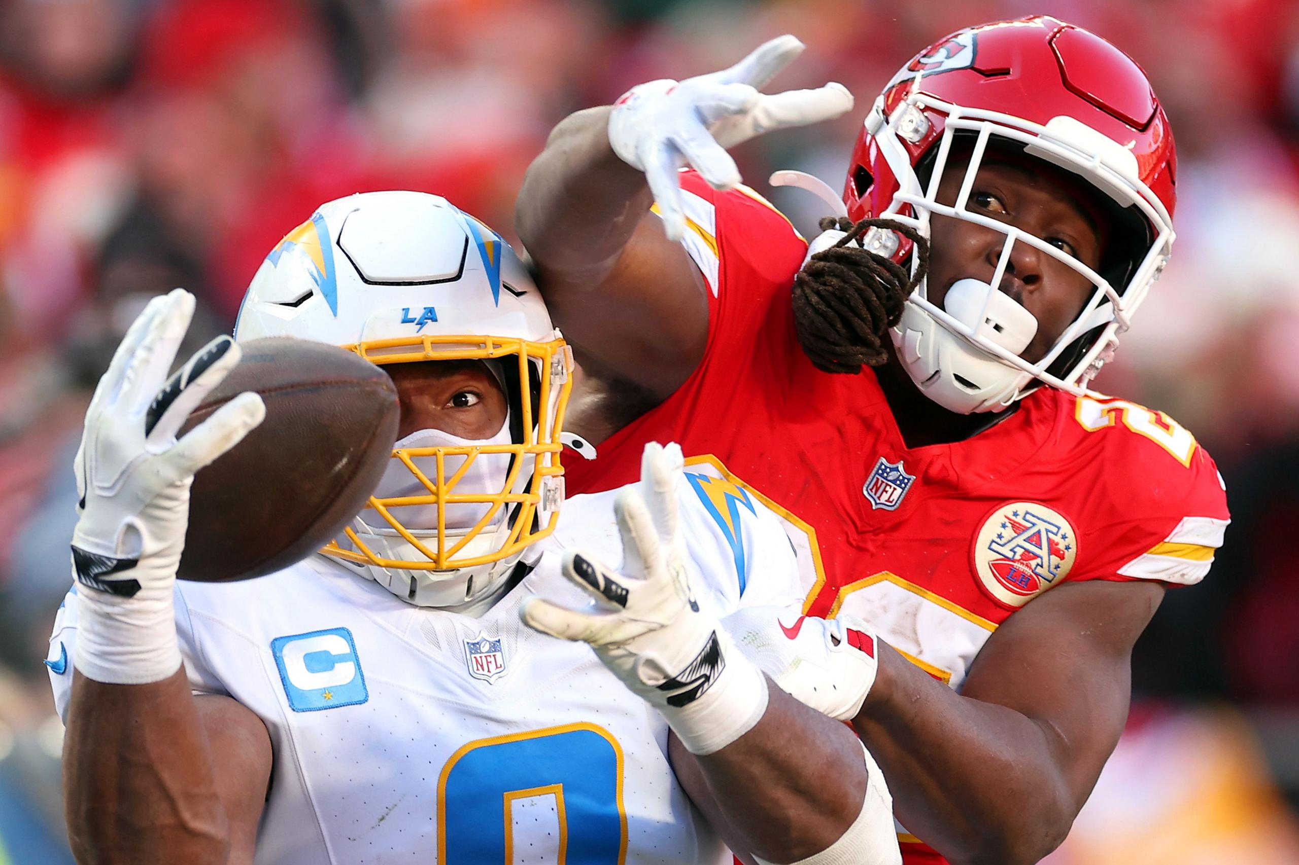 A Kansas City Chiefs defender in a red uniform reaches around a Los Angeles Chargers player in a white uniform, attempting to block or strip the ball during a tightly contested play.