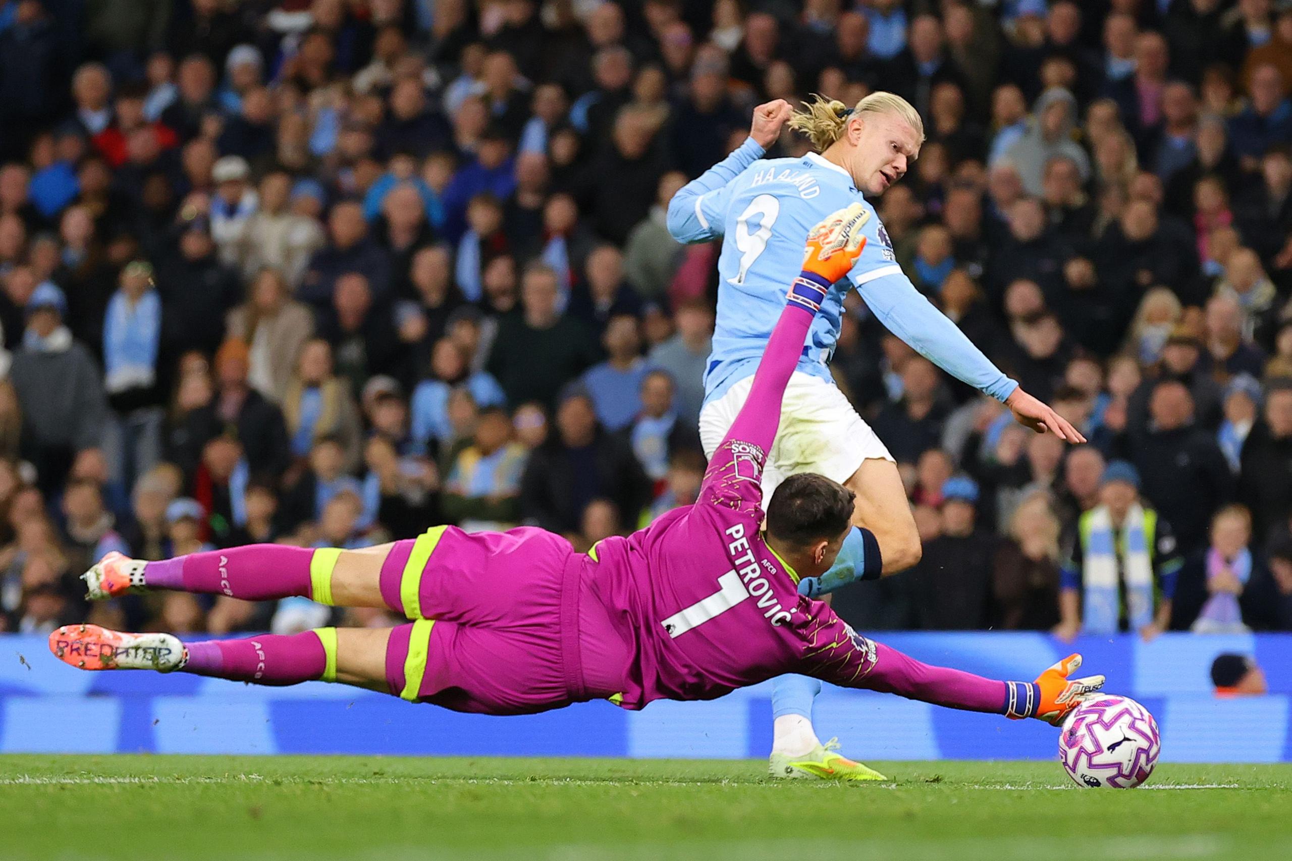 A goalkeeper in a bright pink uniform dives fully extended across the goal area, reaching for a patterned soccer ball. An opposing player in a light blue jersey moves forward with the ball. The background shows a crowded stadium of spectators.