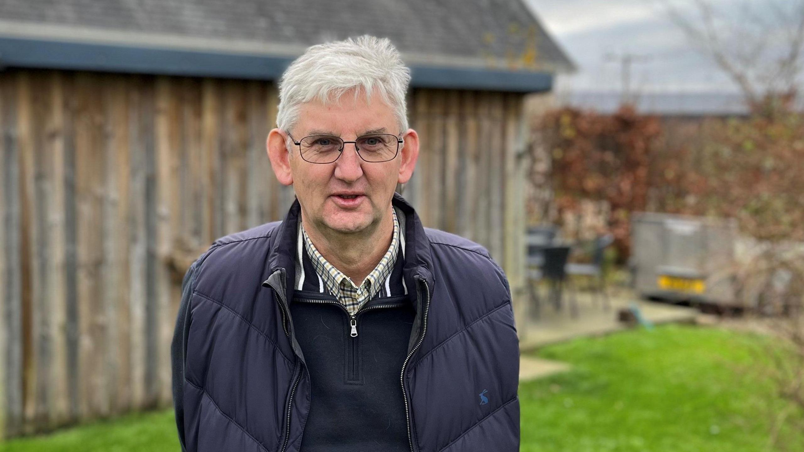 A grey-haired man with glasses, and wearing a blue jacket and blue jumper, standing in front of a garden shed