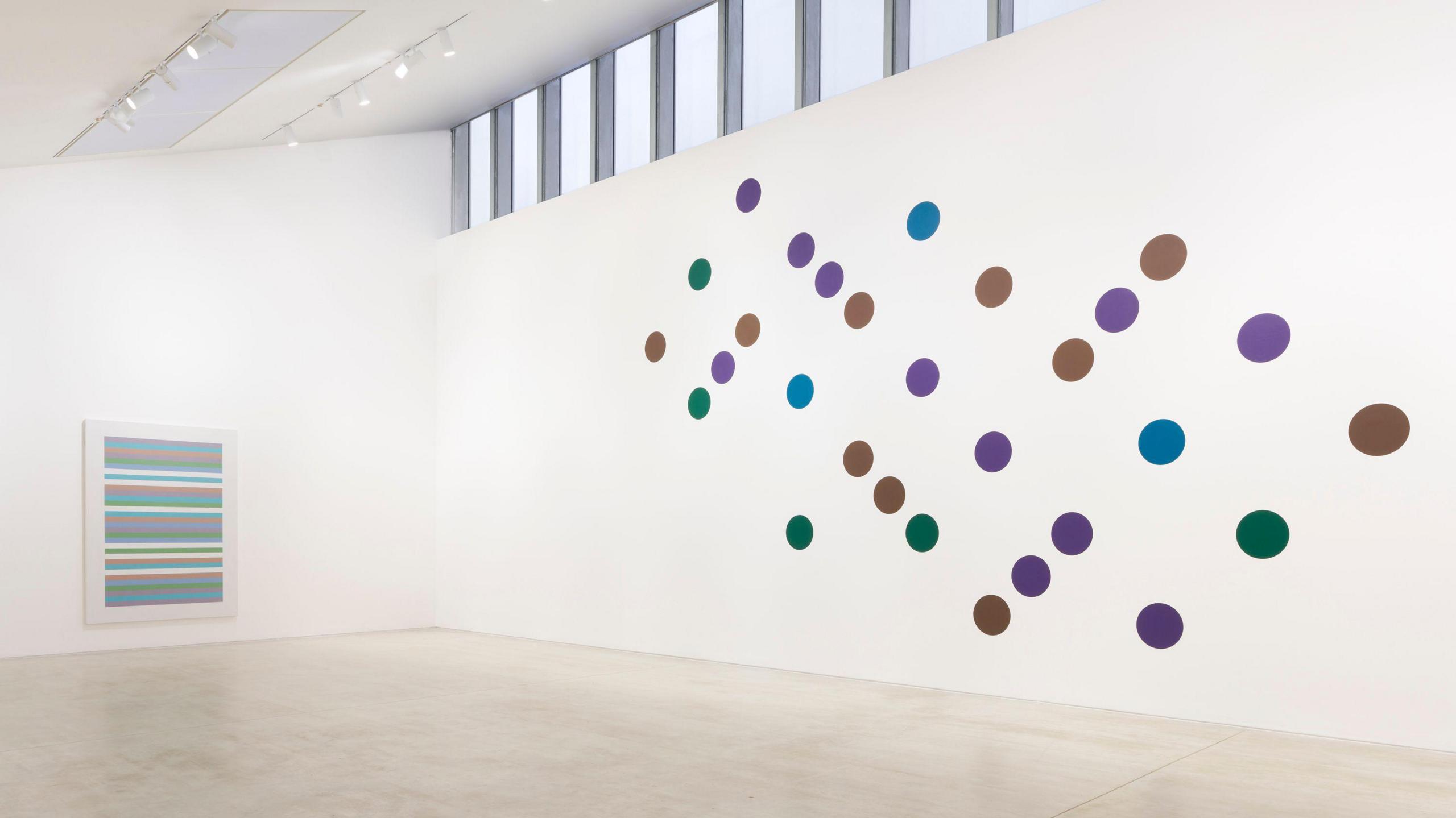 A gallery room with white walls and a row of windows near the roof. On one wall is a large painting consisting of multiple spots in purple, green and brown, seemingly arranged at random but somehow also with a sense of order. On the left is a painting comprised of several horizontal lines in green, pink, blue, white and some other colours.