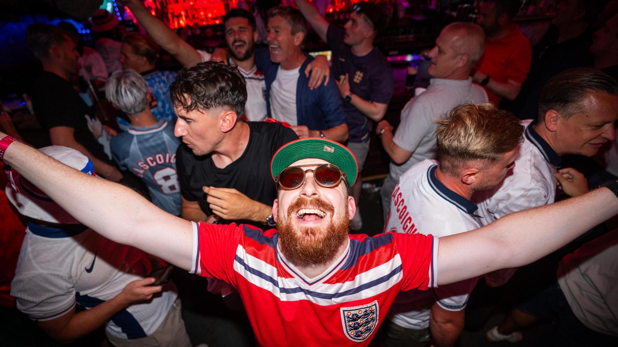 An England fan wearing a red shirt and sunglasses puts his arms out in celebration in a packed bar in Boston ahead of the game against Ghana