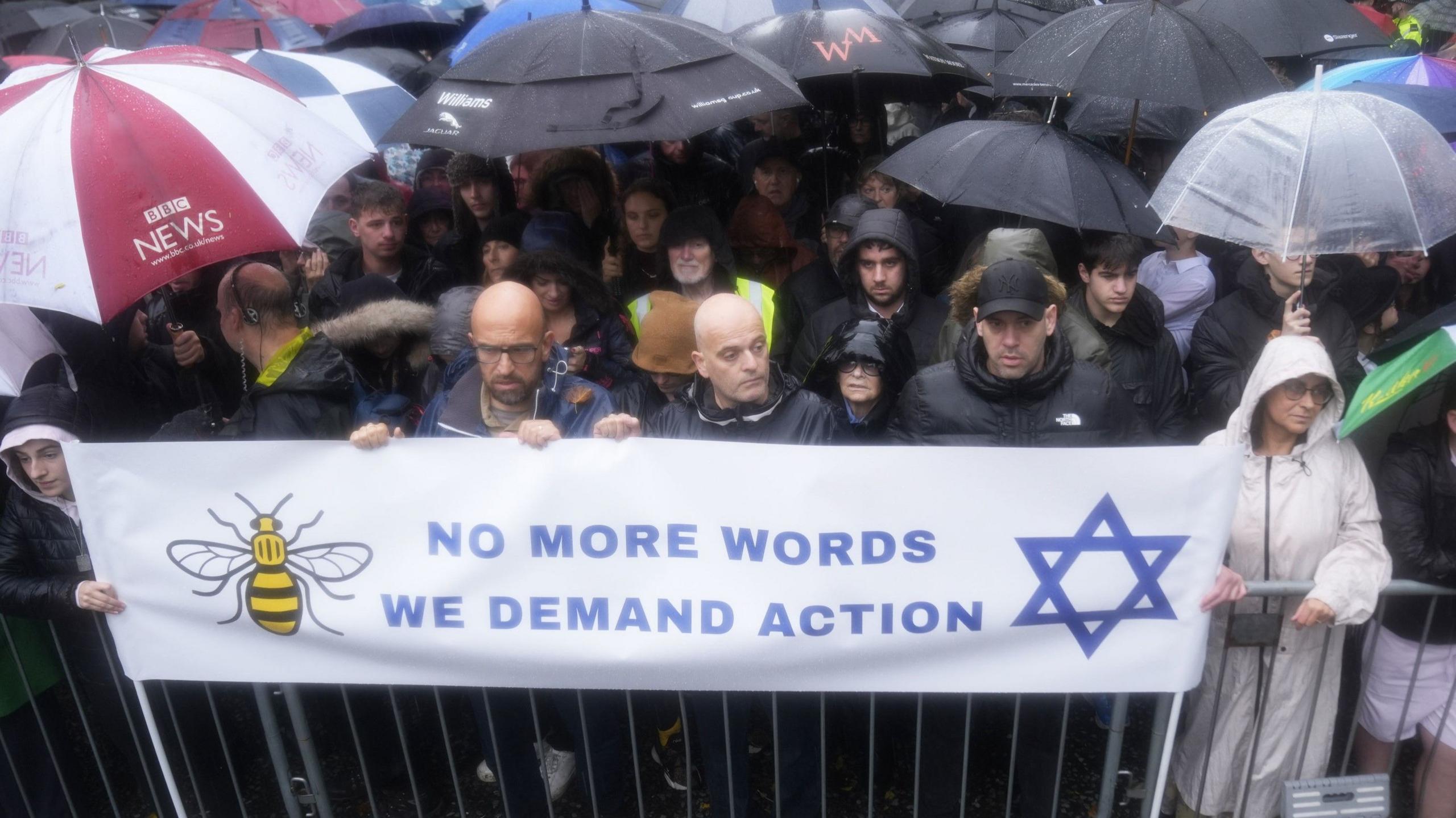 A large group of people standing behind railings and a banner that says 'No more words, we deman action', with a picture of a bee and a Star of David. People look serious and many are carrying umbrellas as it is raining heavily.