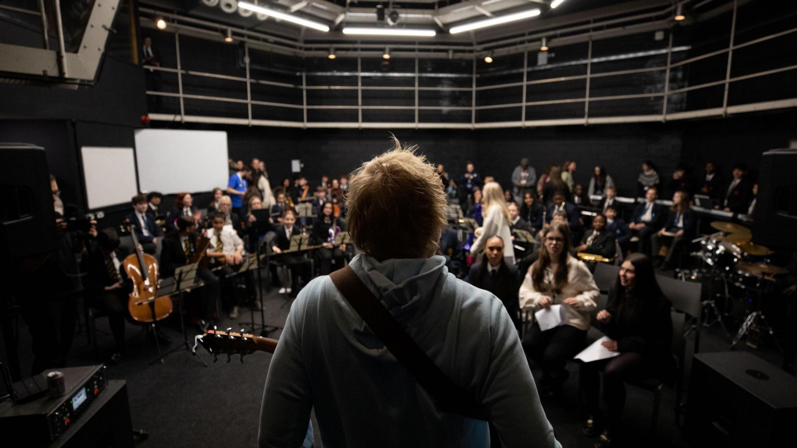 Ed Sheeran with his back to camera, with his guitar strapped to him, in a large music room with students in front of him holding a variety of instruments