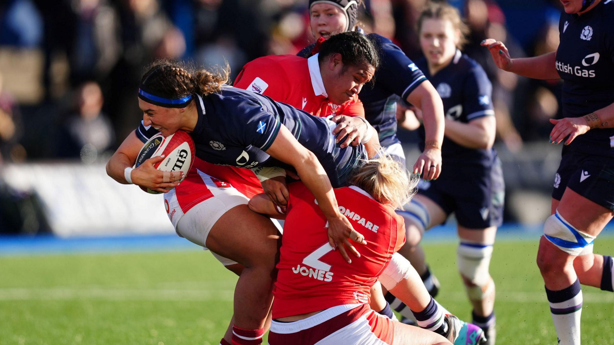 Emma Wassell is tackled by two Welsh players during a Six Nations match in 2024. She is reaching out with the ball in one hand, in a blue kit, while the Wales players are grabbing her while dressed in red and white.