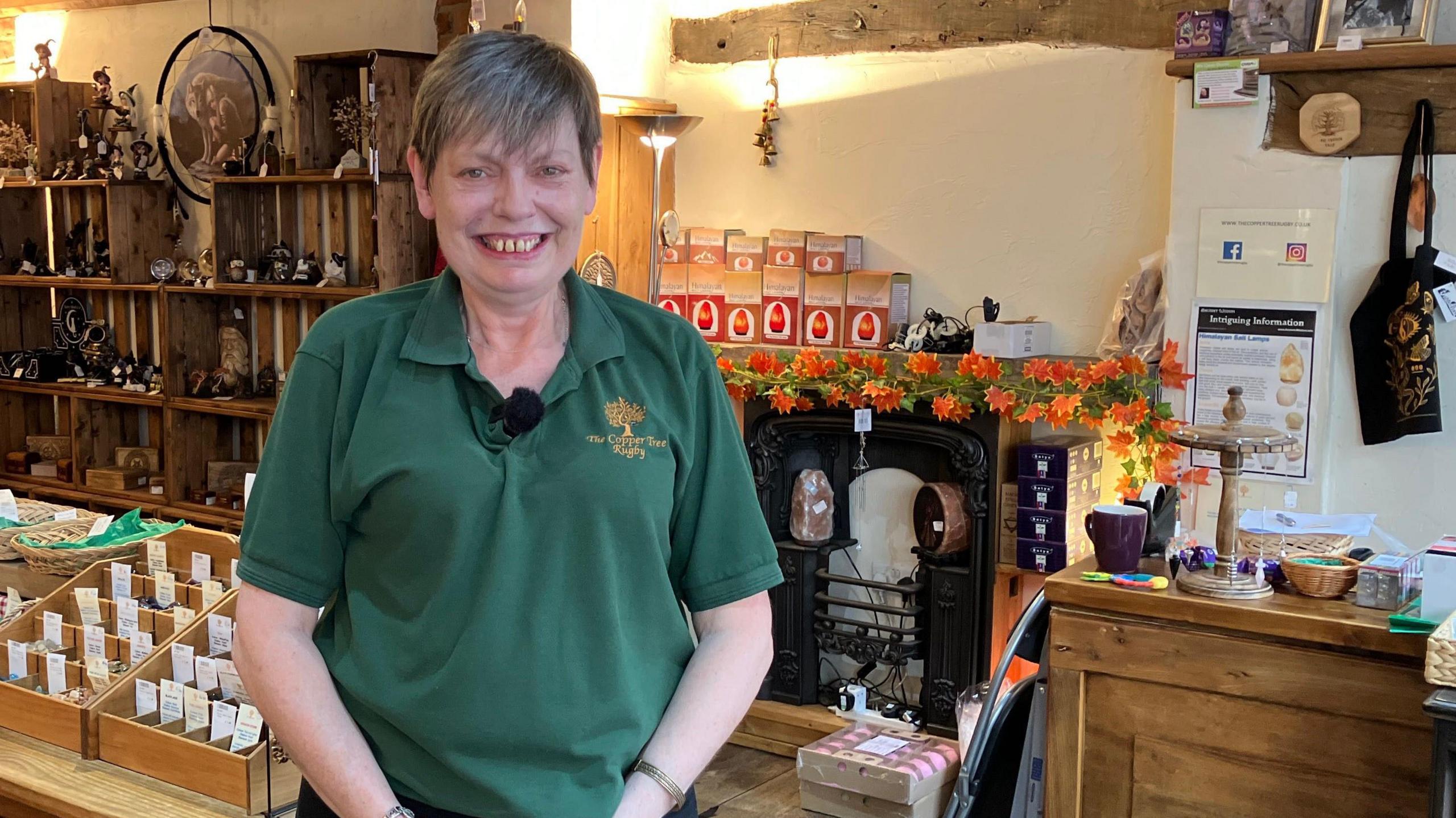 Helen Simmonds, stands in front of the counter in her gift shop. She is wearing a green top with the words The Copper Tree Rugby on it. There are shelves of gifts including ornaments, dream catchers and baskets of products. The shop is lit with bright lights.