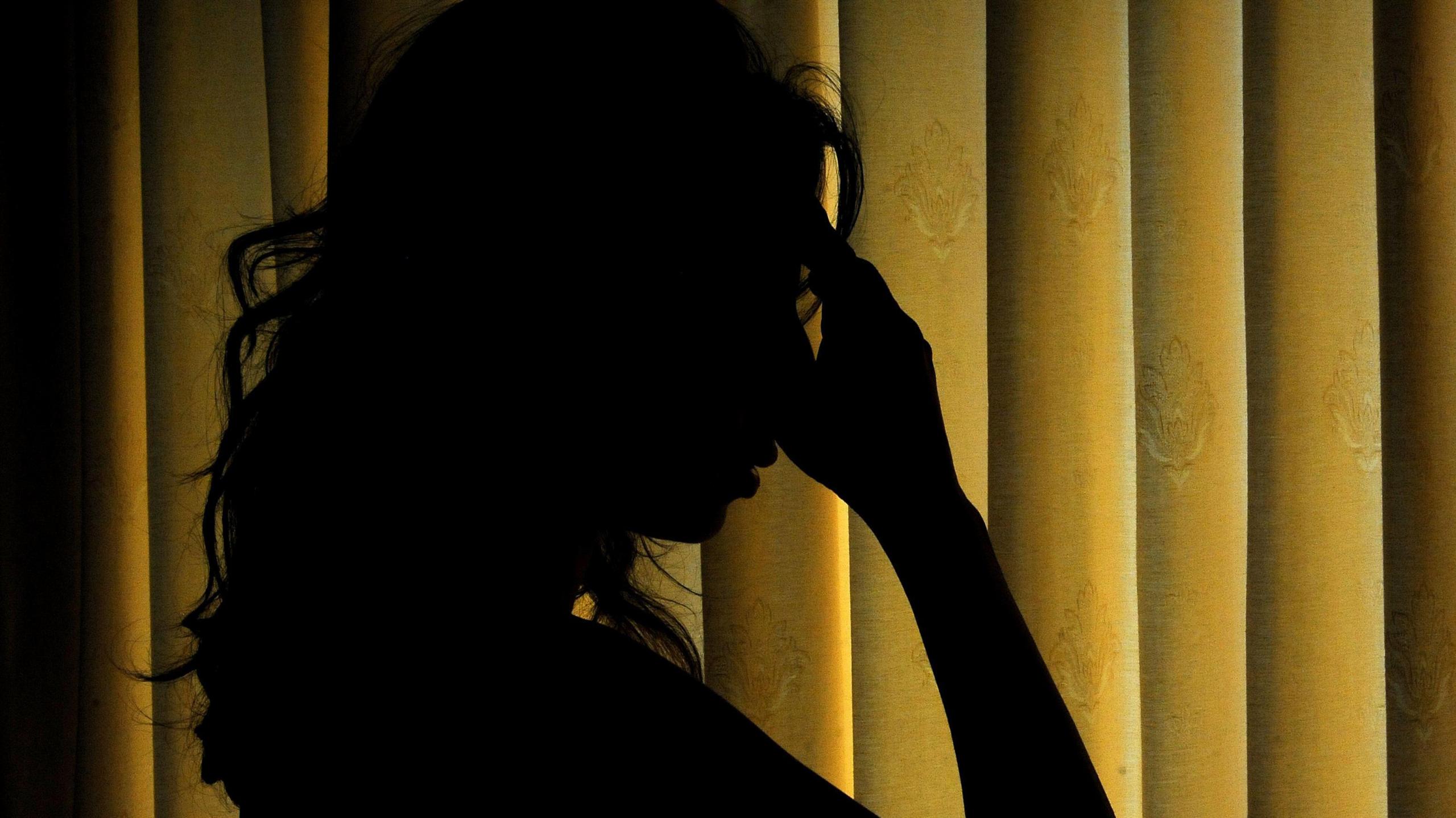 The silhouetted woman with long hair and her hand to her head in front of curtains