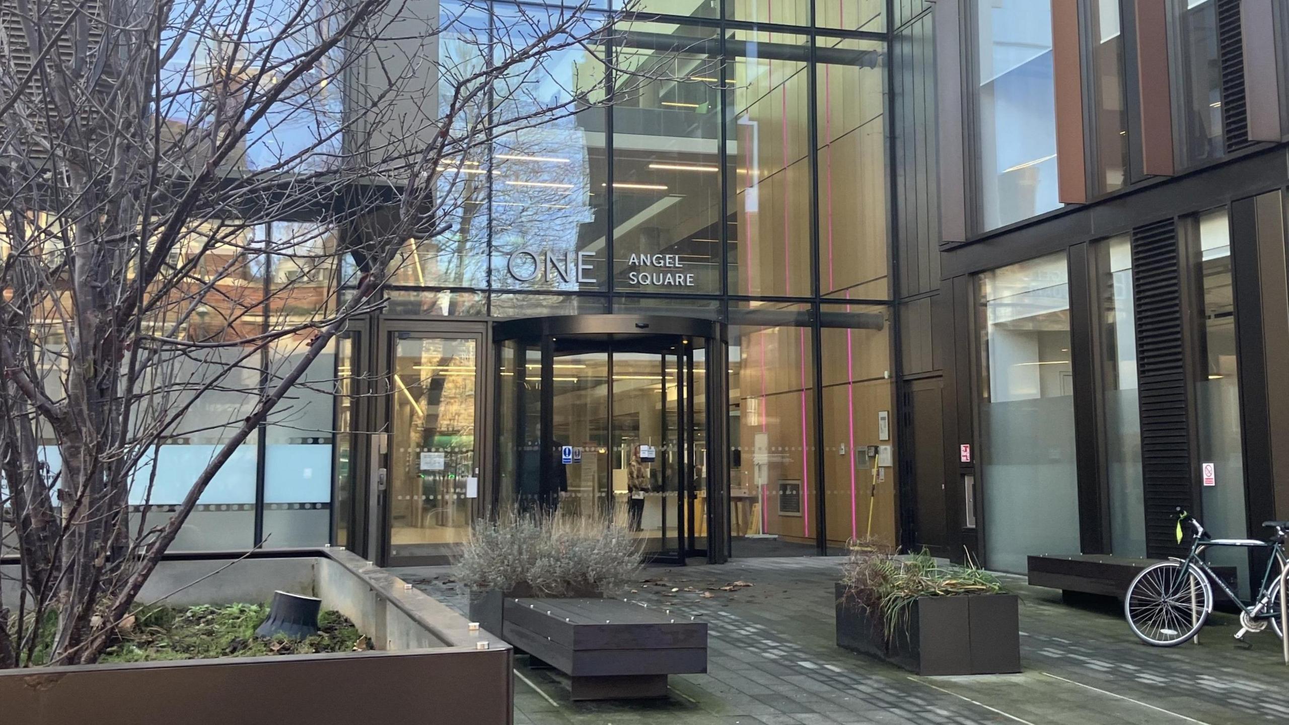 The entrance to West Northamptonshire Council's headquarters. It is a large building made up of clear glass panels. A revolving door can be seen as well as some benches, a bike and planted areas.