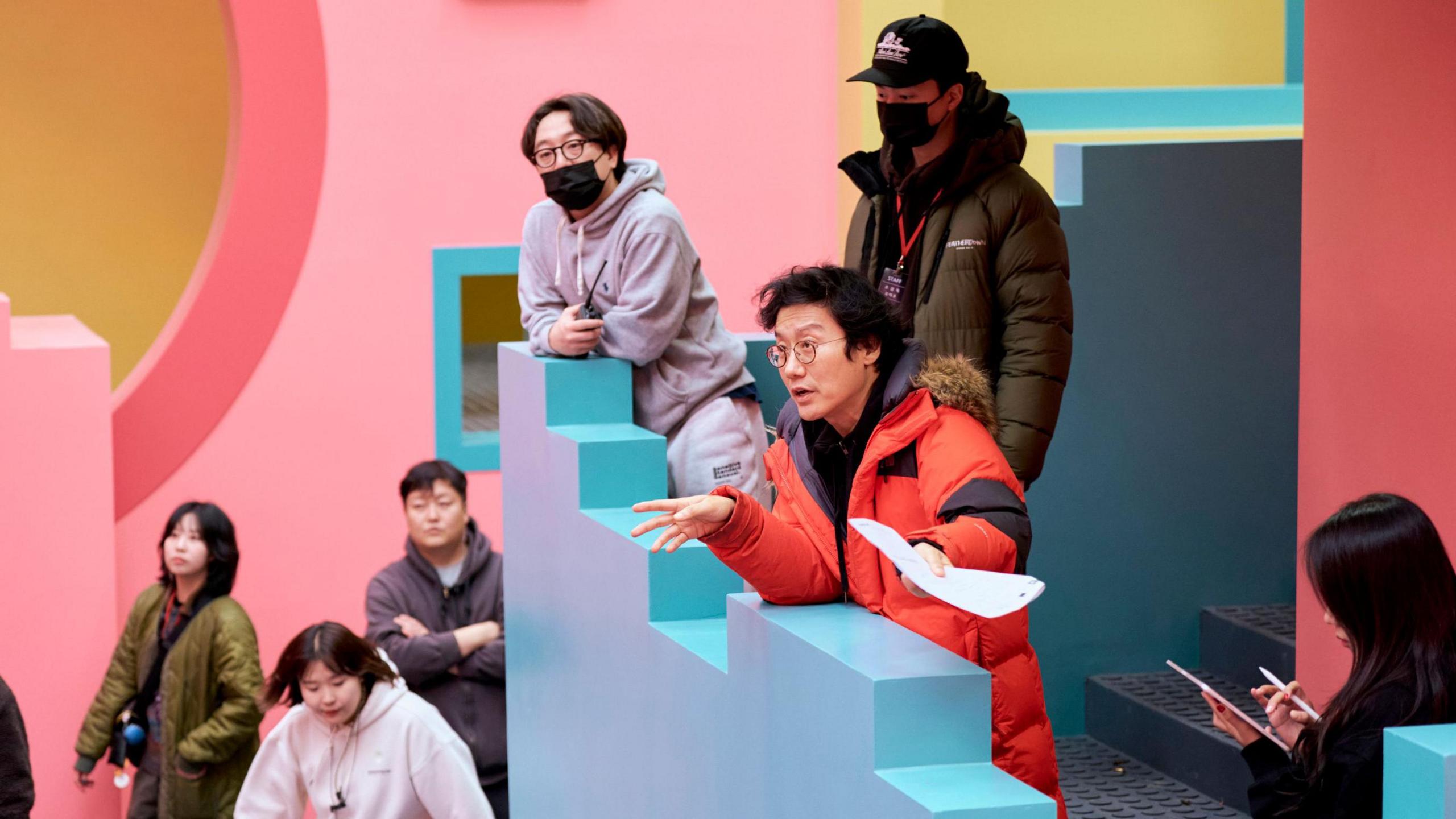 Hwang in an orange jacket on the set for Squid Game 2, adressing staff on the distinctive  staircase which is painted blue and pink  