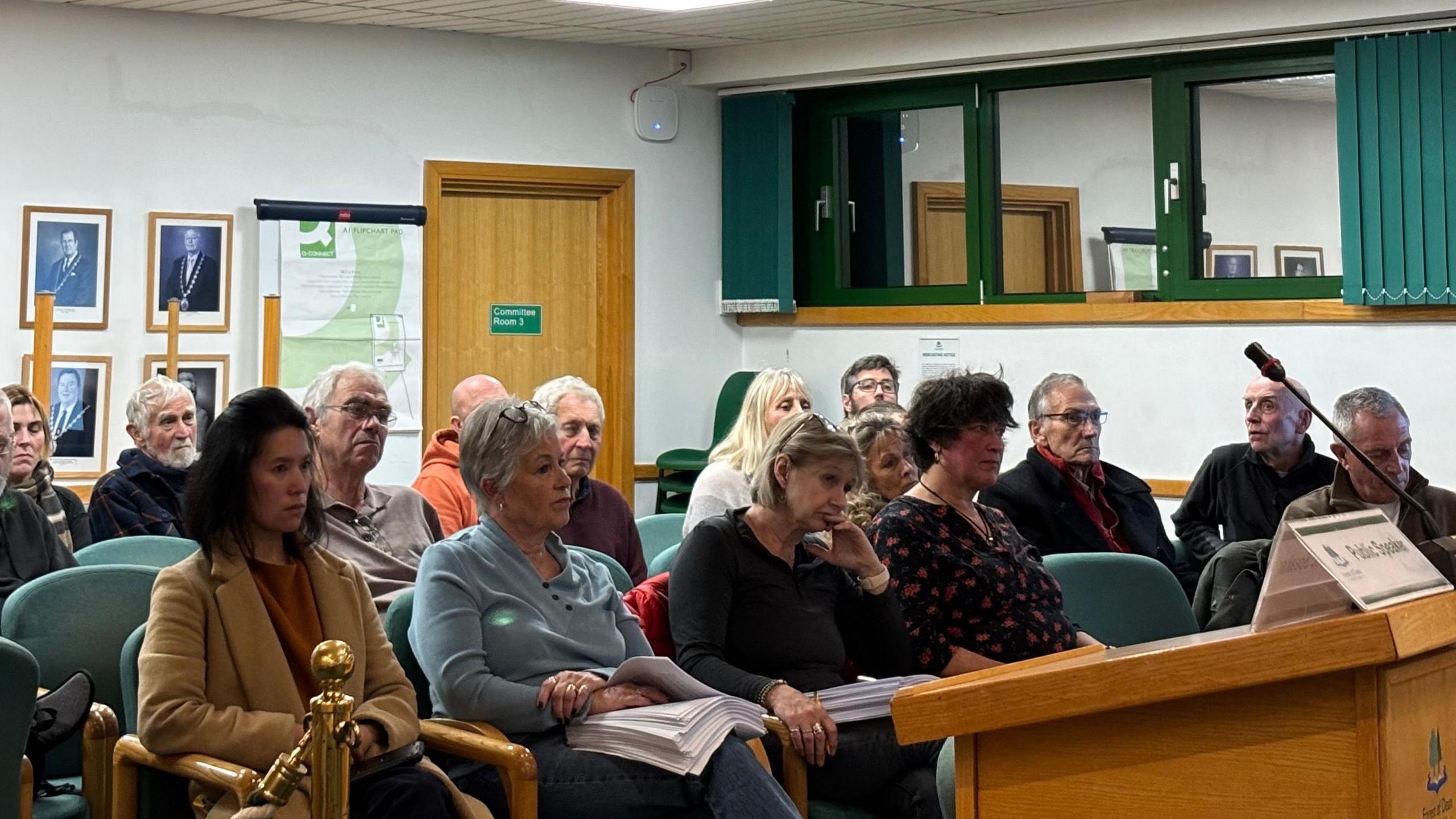 Members of the public sitting a listening to councillors speak in the Forest of Dean District Council offices in Coleford. More than a dozen people are visible and listening to the debate. There is a lectern with a mic on top of it at the front of the image.