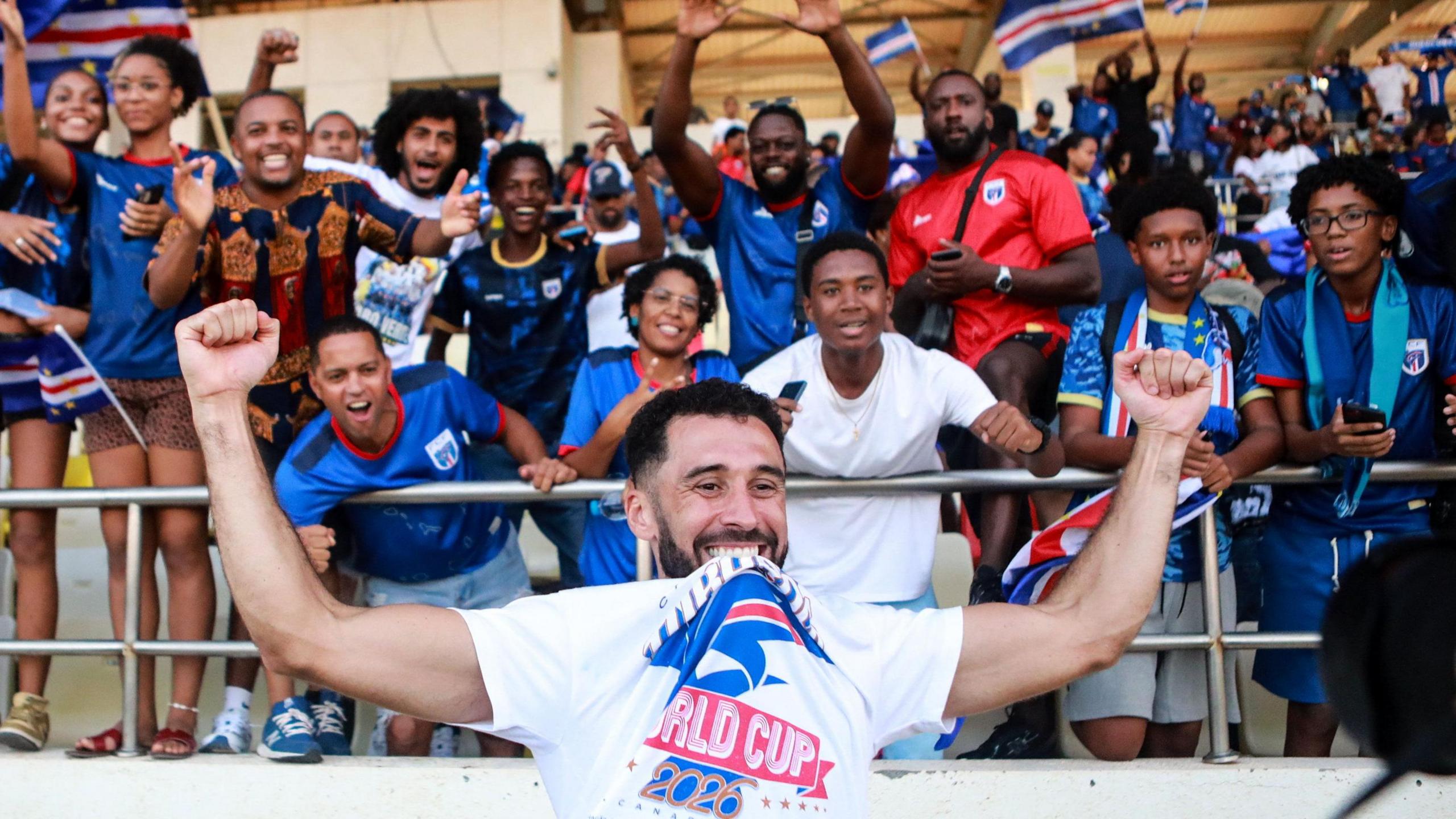 Roberto Lopes, visible from chest up in the foreground of the image, holds his arms up high as he celebrates Cape Verde qualifying for the 2026 World Cup. He wears a white T-shirt with the text World Cup 2026 on it, which he raises up by biting it between his teeth. Twelve joyful Cape Verde fans, mostly wearing blue team jerseys, are seen in the stands behind him, with other people and some Cape Verde flags seen out of focus in the distance