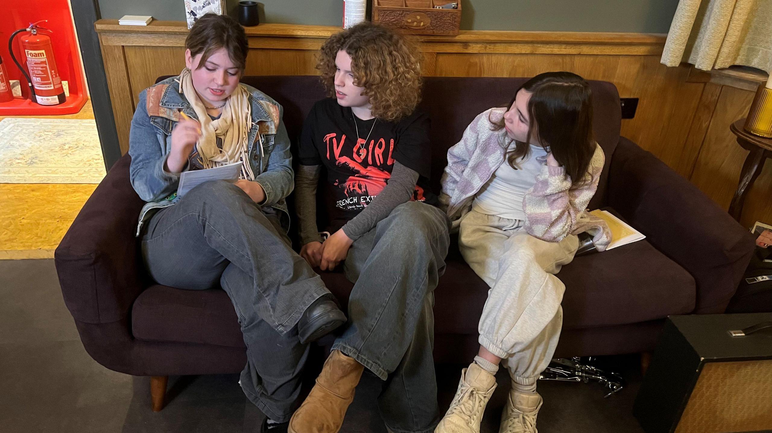 Three young women sit on a dark brown sofa inside a recording studio. One of them is writing on a notepad and the other two are looking at her. All three are dressed in casual clothing