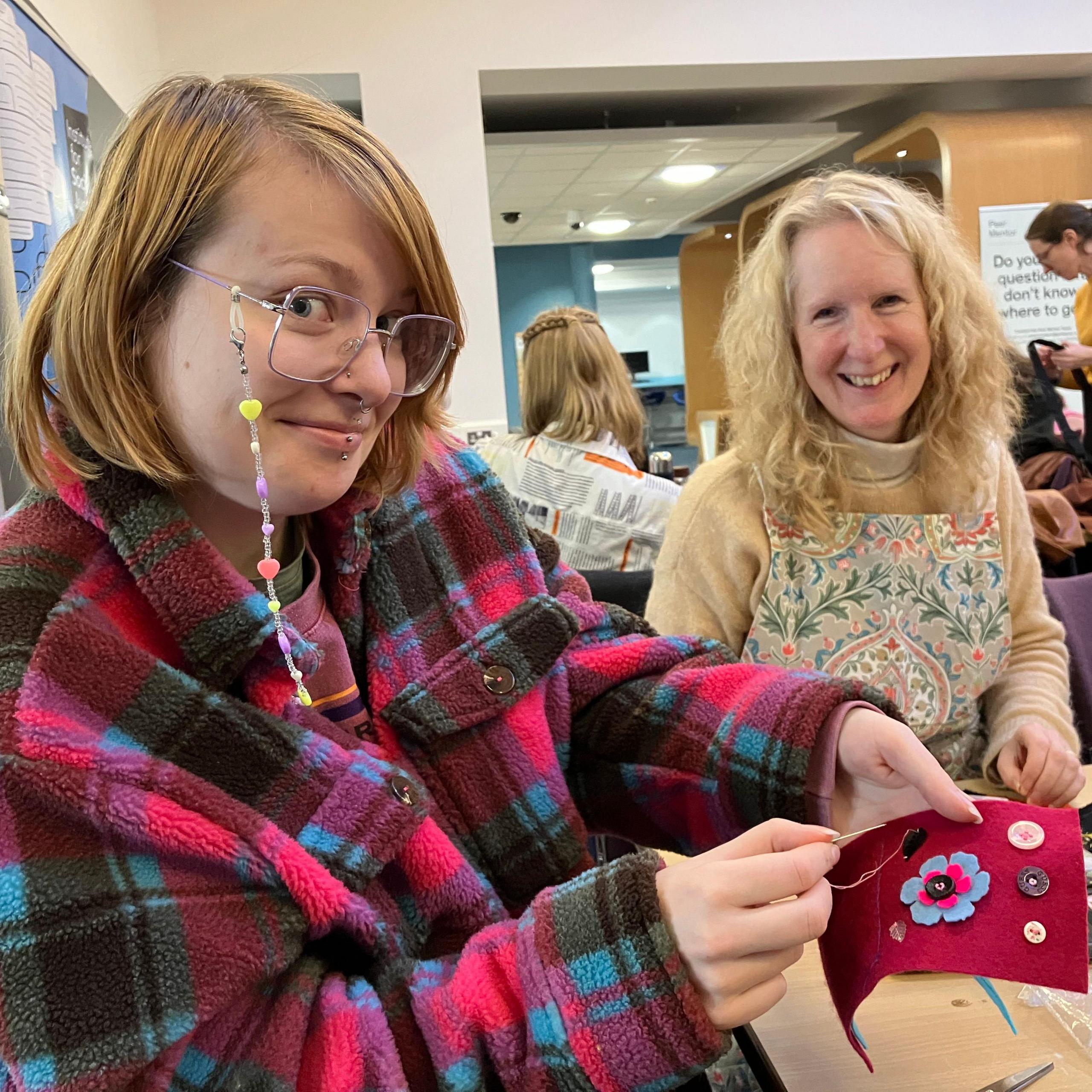 Student Bailee Wray holds up a piece of cloth with buttons sewn onto it, while Tadcrafters founder Su Morgan looks on.