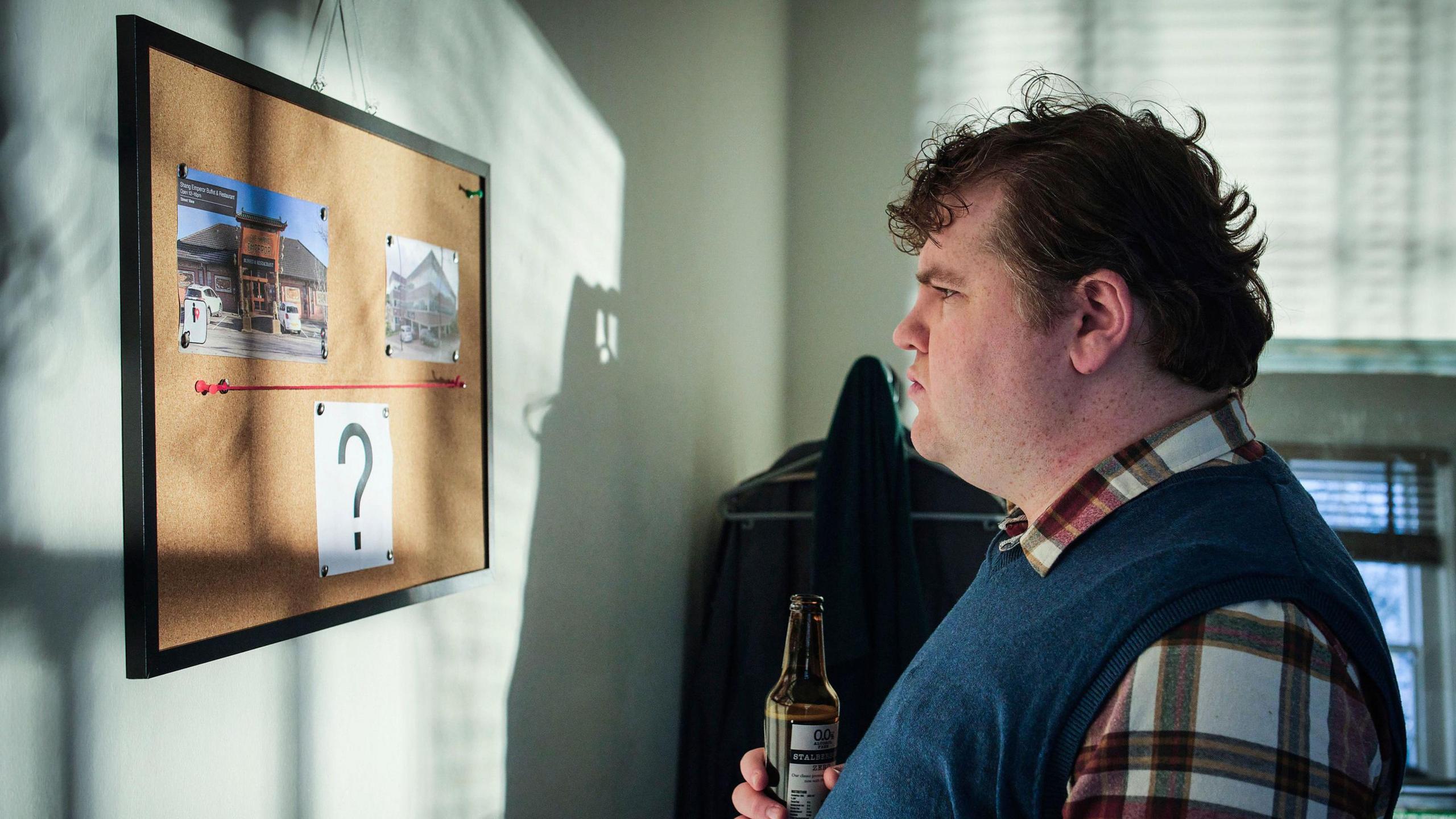 Ethan Lawrence as Ben in Boat Story. Ben is pictured off duty, holding a beer and looking at a cork board with two pictures connected by a red thread with a question mark printed underneath. Ben is a man in his 30s with curly brown hair and wears a blue vest over a red and white plaid shirt 