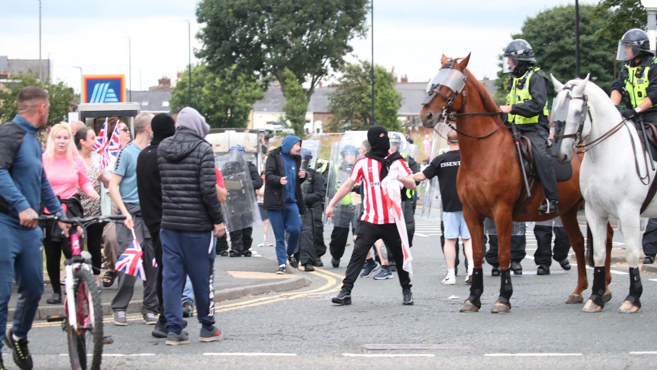 People look on as man in Sunderland shirt and balaclava talks to police officers on horses wearing riot gear