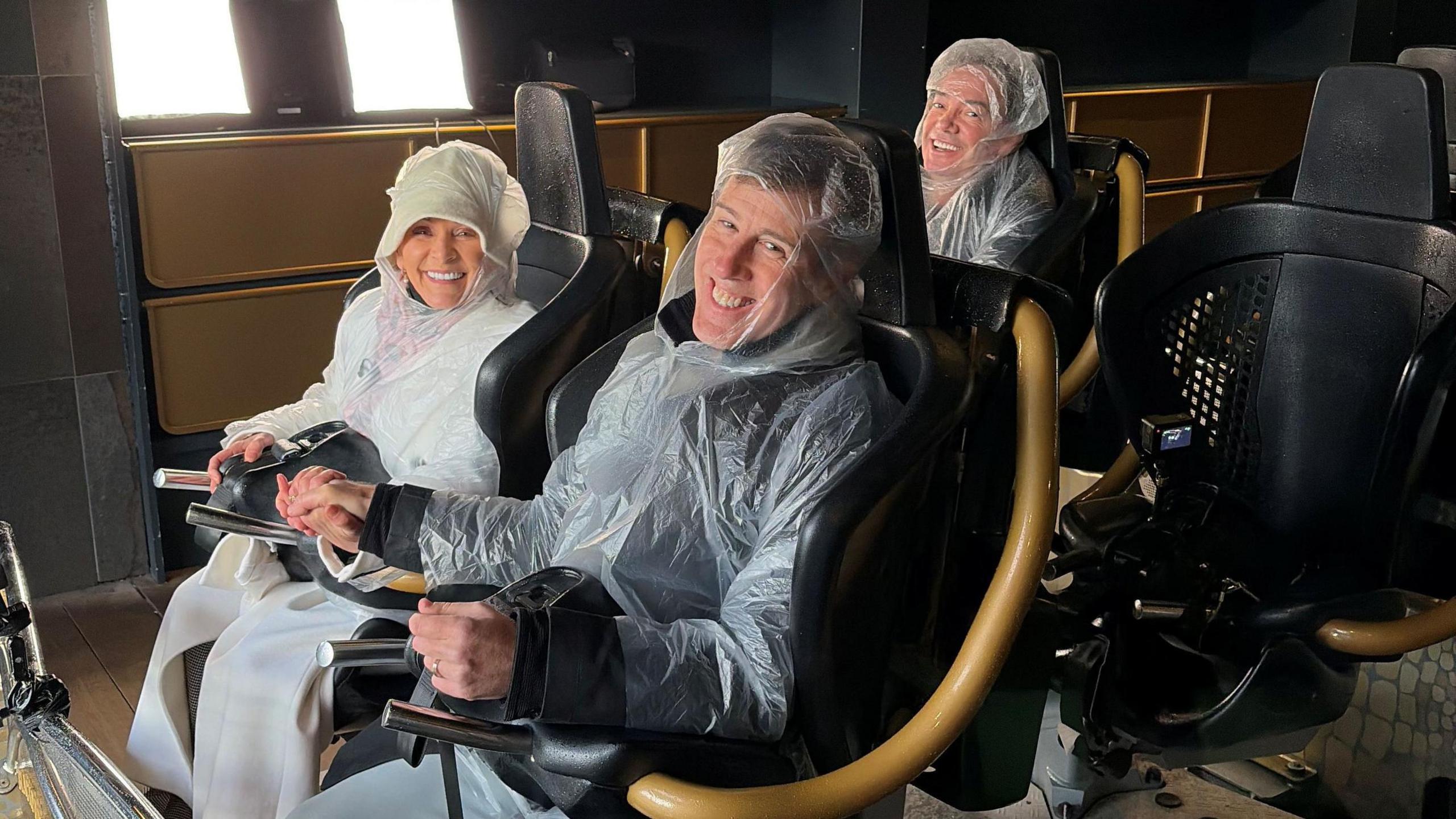 Shirley and Anton sit in the front seats of a rollercoaster while Craig sits behind. They grin as they wear rain macs.