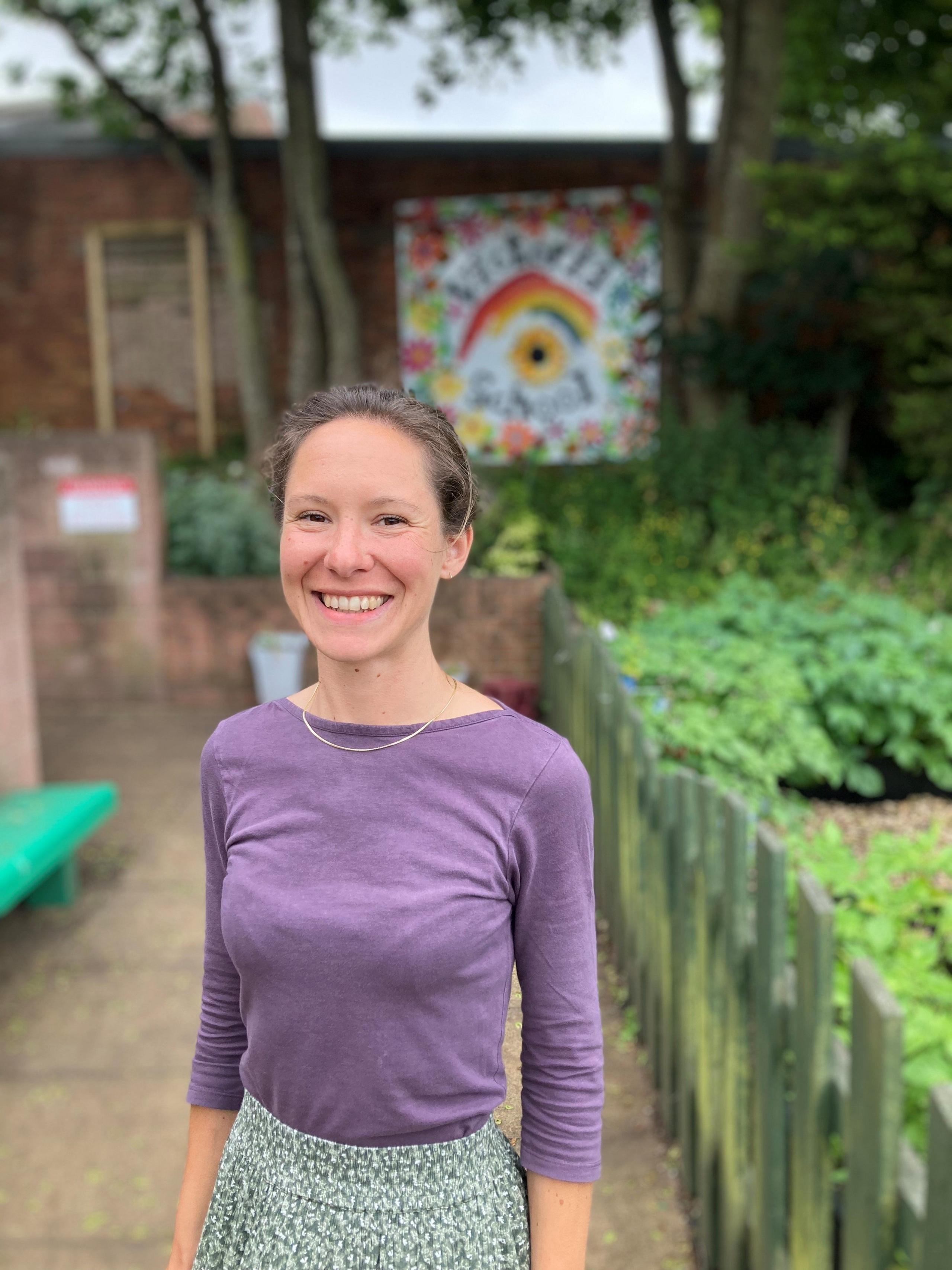 Poet Melissa Davies smiling in the schools garden. She's wearing a purple top.  A piece of art with a rainbow on it can be seen in the background.