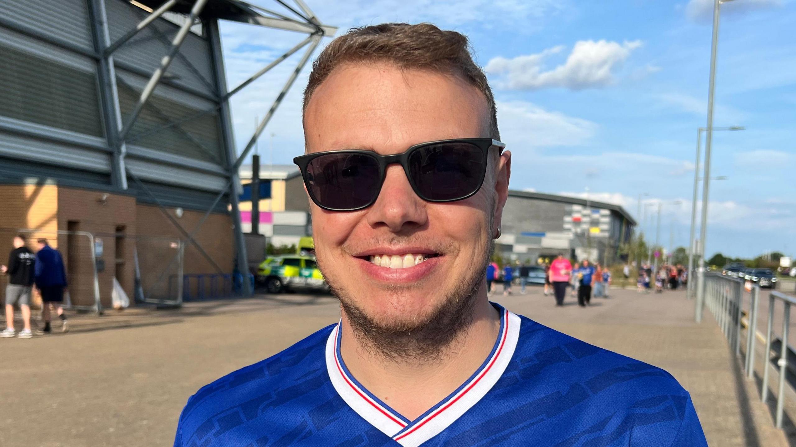 Lee Pike is stood in the middle of the picture smiling at the camera. He has short brown hair and stubble, and is wearing black sunglasses and a blue Ipswich Town football T-shirt. Behind him is part of the stadium on the left and a road on the right.