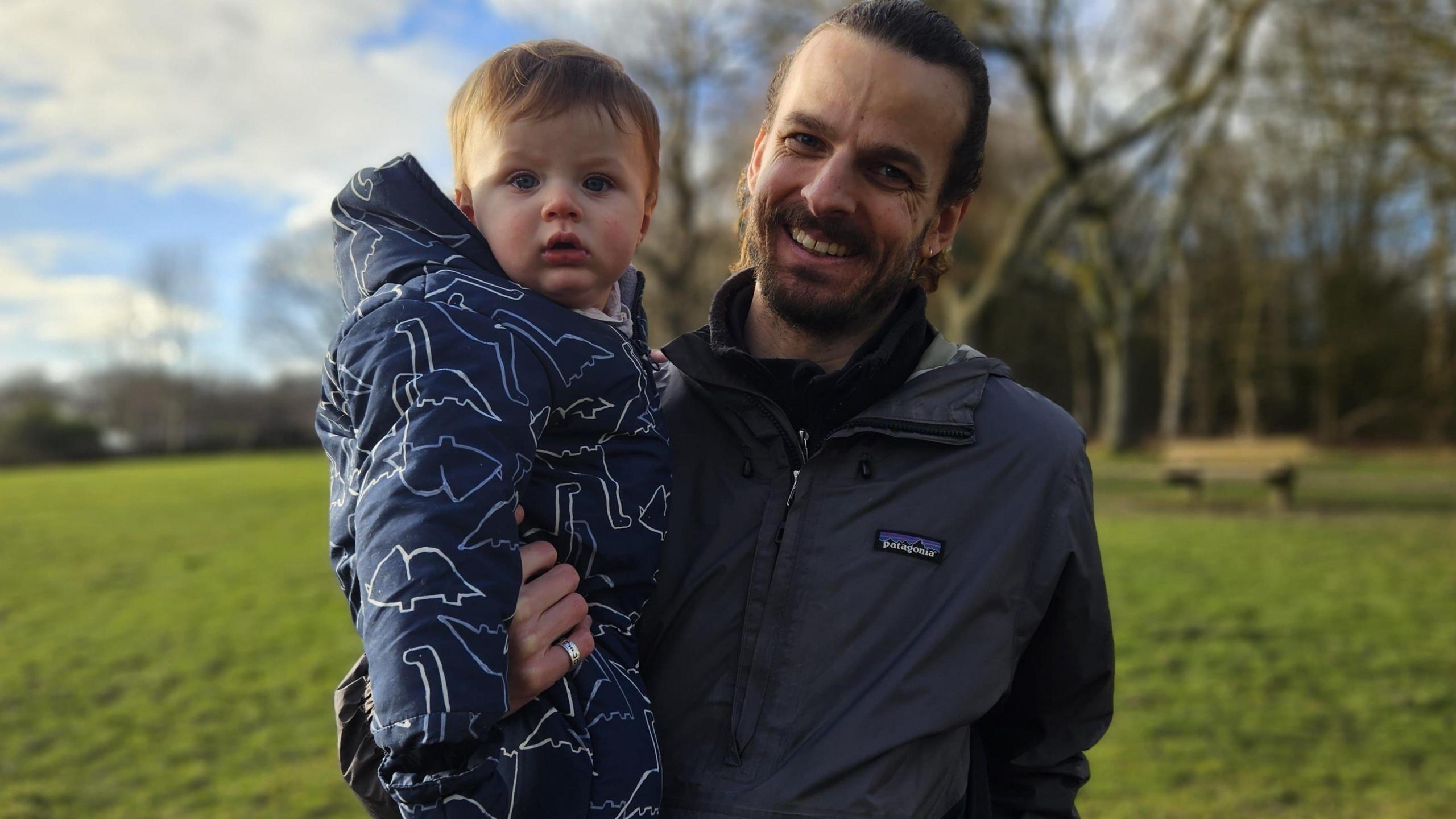 A smiling young man in his 20s or 30s, brown haired and with a short beard, stands in the middle of a field. He is holding a baby boy who is wearing a blue patterned puffy romper suit.