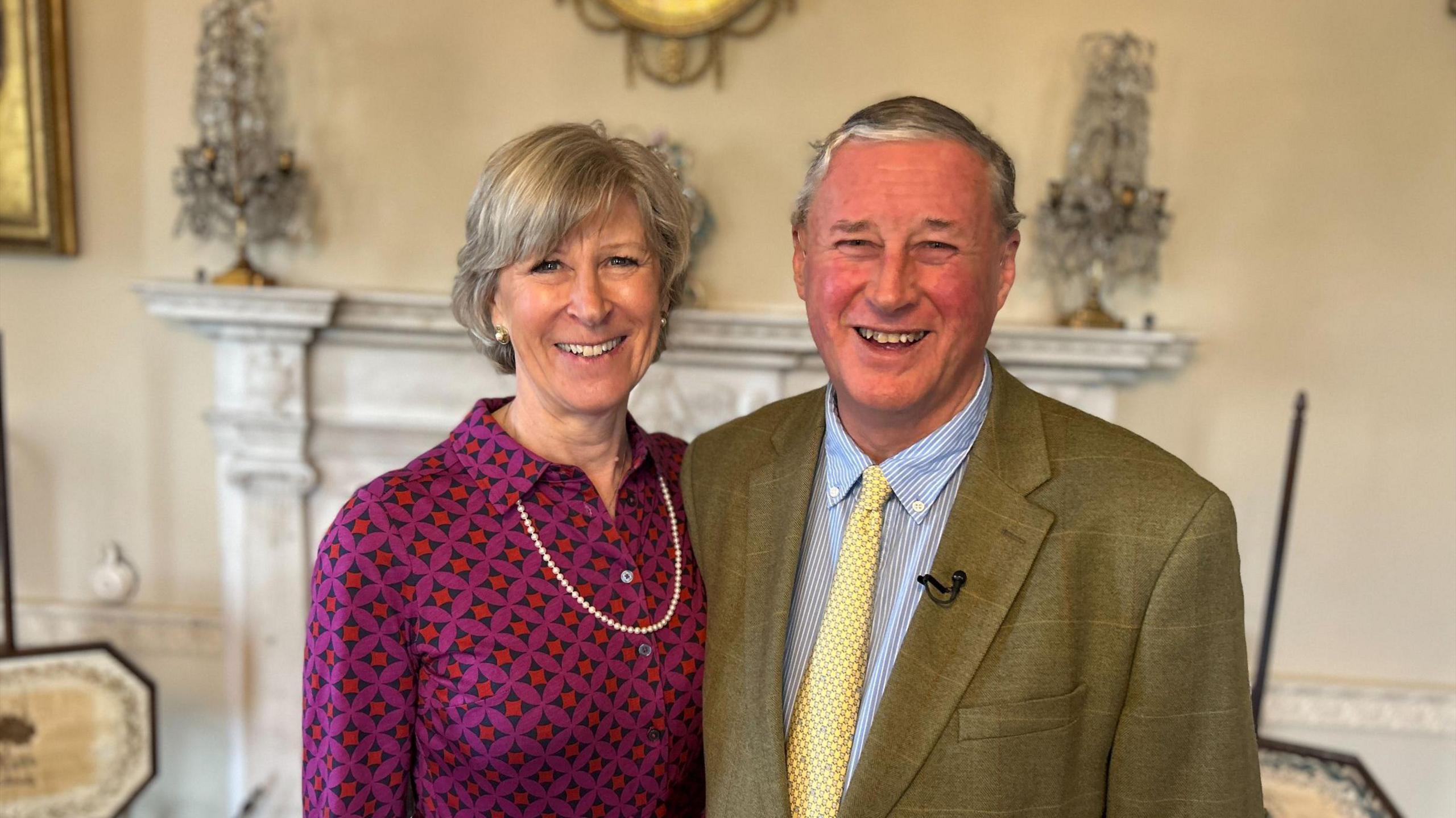 A woman with short hair wearing a purple and red blouse with a single string of pearls arm around a man wearing a green and brown jacket with a blue shirt and pale yellow tie