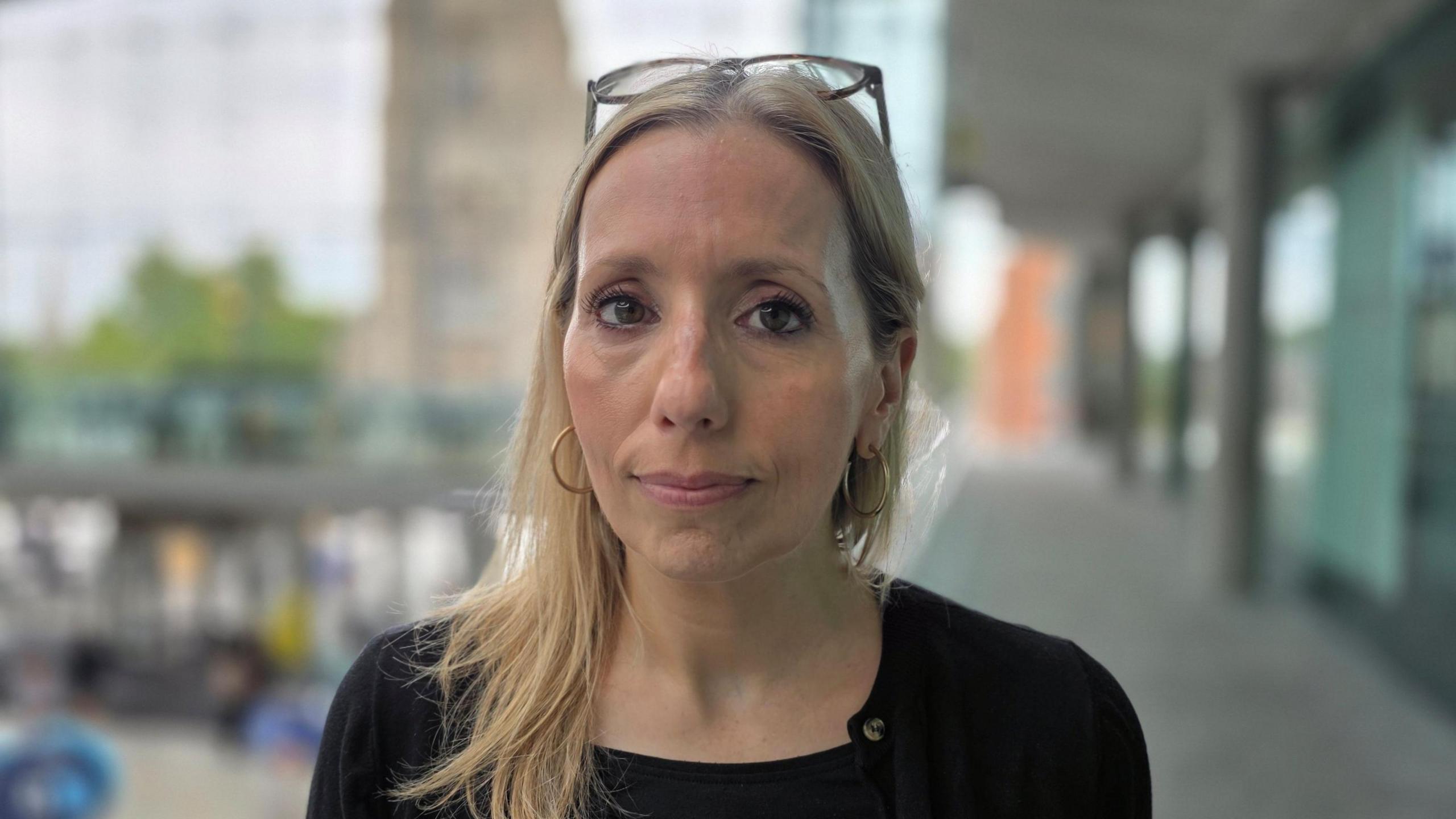 Sarah Taylor is wearing a black top. Her glasses are placed on top of her head. She is standing inside Norwich's Forum building, and a church tower is in the very background the other side of the glass frontage, out of focus.