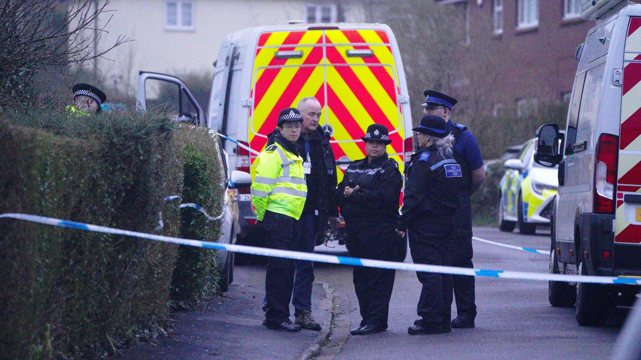 A group of police officers standing behind a police van. They are all in black uniforms and one of them is wearing a yellow high vis jacket. Two of the officers are looking forward, towards the camera, while the others are looking at each other. 
