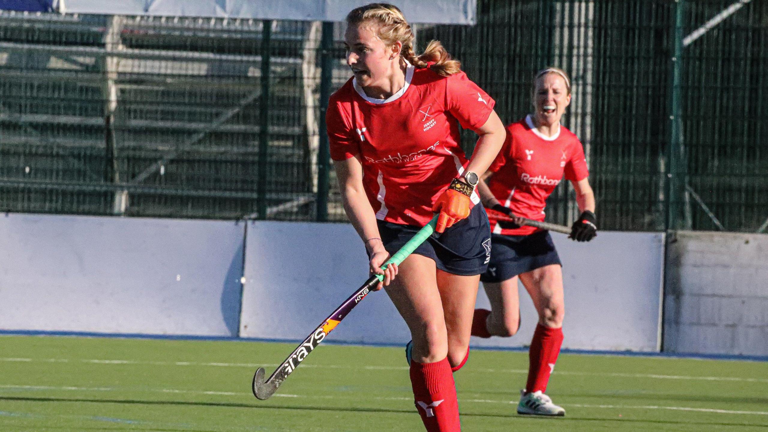 Two women are playing hockey wearing red shirts, blue shorts and red socks