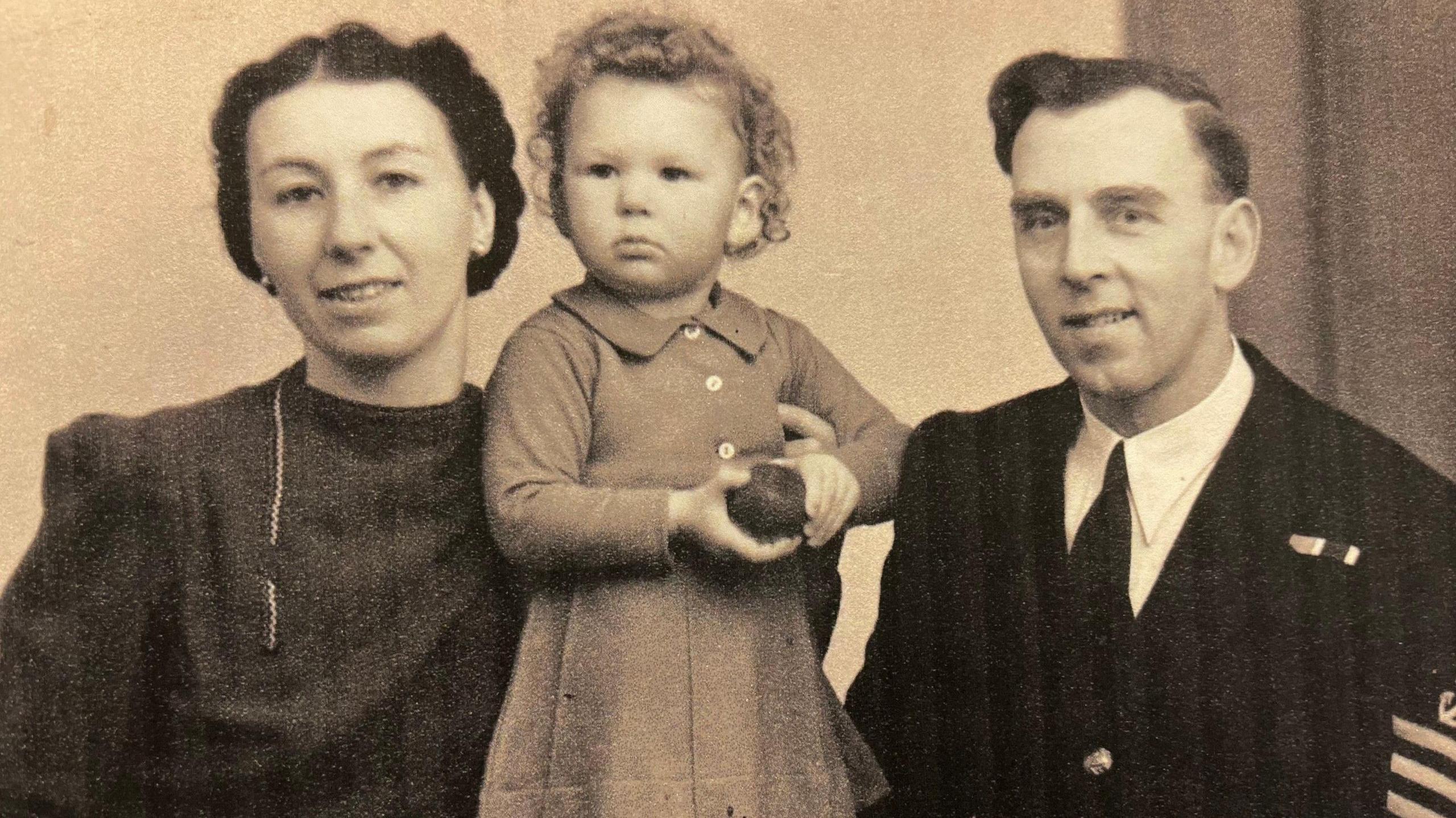 A sepia photograph of a young couple - Margery and Reginald Arliss - with their toddler daughter Judy stood between them. The photo is believed to have been taken in late 1943. Margery is wearing a high-necked dark coloured dress, with her hair pinned up. A young Judy - around 18 months old - is wearing a lighter coloured dress with long sleeves and appears to be holding a toy ball. Her father, Reginald, is wearing his Navy uniform - it's a dark blazer with three stripes on his left shoulder, and medals pinned to his left breast pocket. He's also wearing a white collared shirt and a dark tie. The parents are smiling and looking at the camera. 