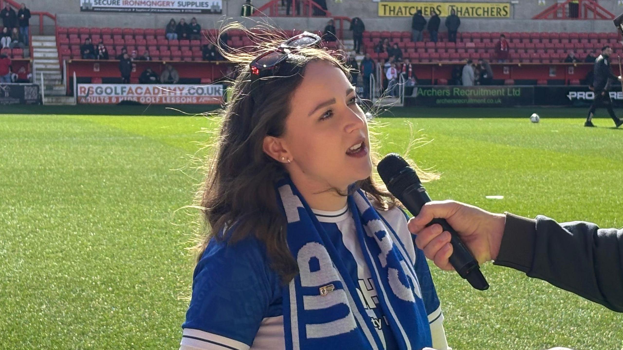 A young woman is interviewed on a football pitch. She is wearing a blue and white Bristol Rovers scarf and jersey, and has long brown hair. She has sunglasses perched on top of her head. The arm of a man can be seen holding a microphone to her mouth.