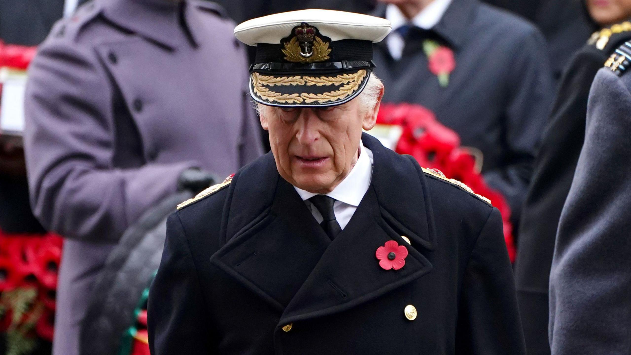 King Charles in Royal Navy uniform during the Remembrance Sunday service at the Cenotaph in London. He is wearing a white Royal Navy officer hat, and a dark coat, with a poppy on the left lapel.