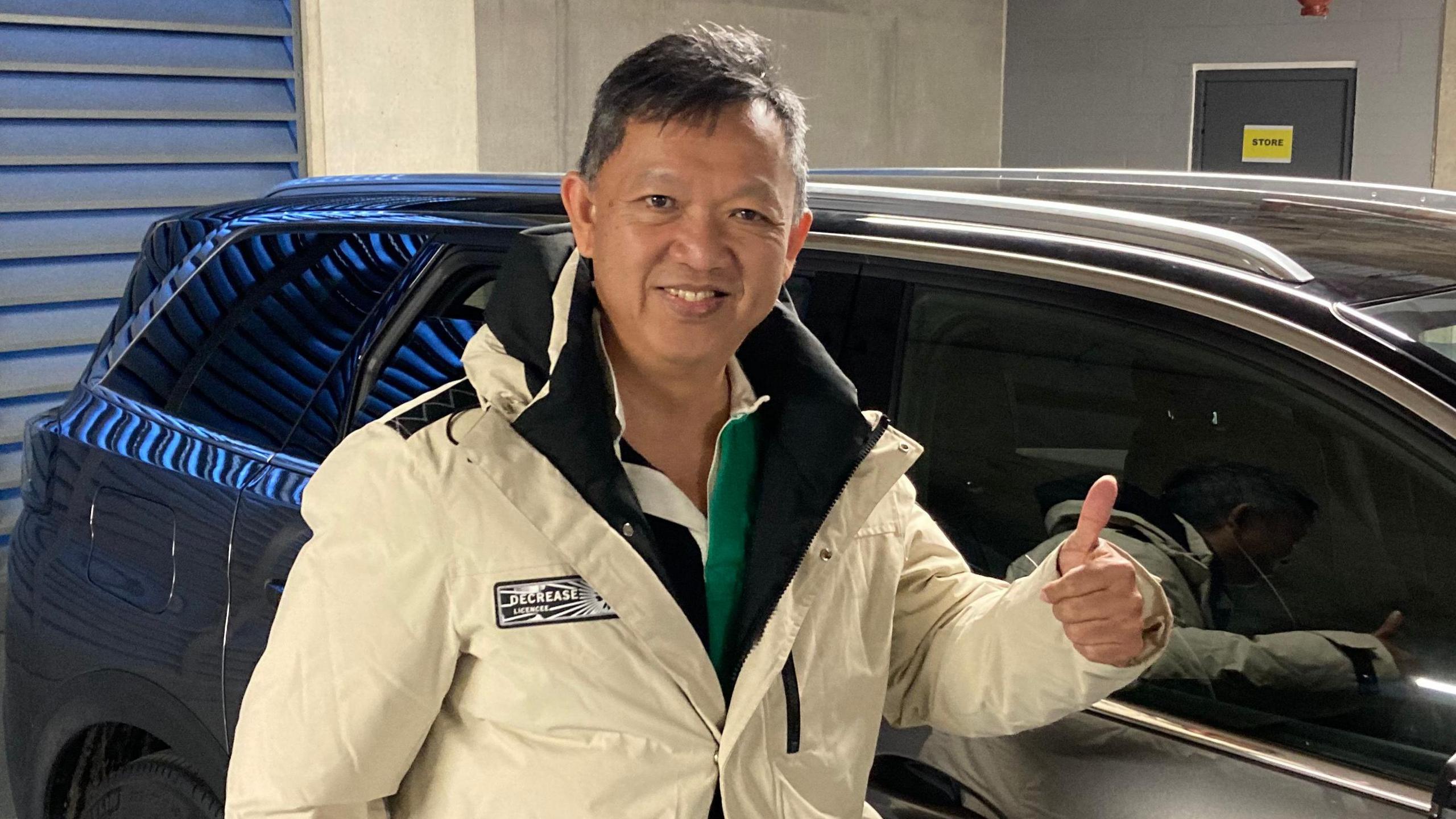 Ling Yohoya in a cream off white coloured coat, he is looking at the camera and smiling and is giving the camera a thumbs up. He is stood next to a large grey car in a underground car park.